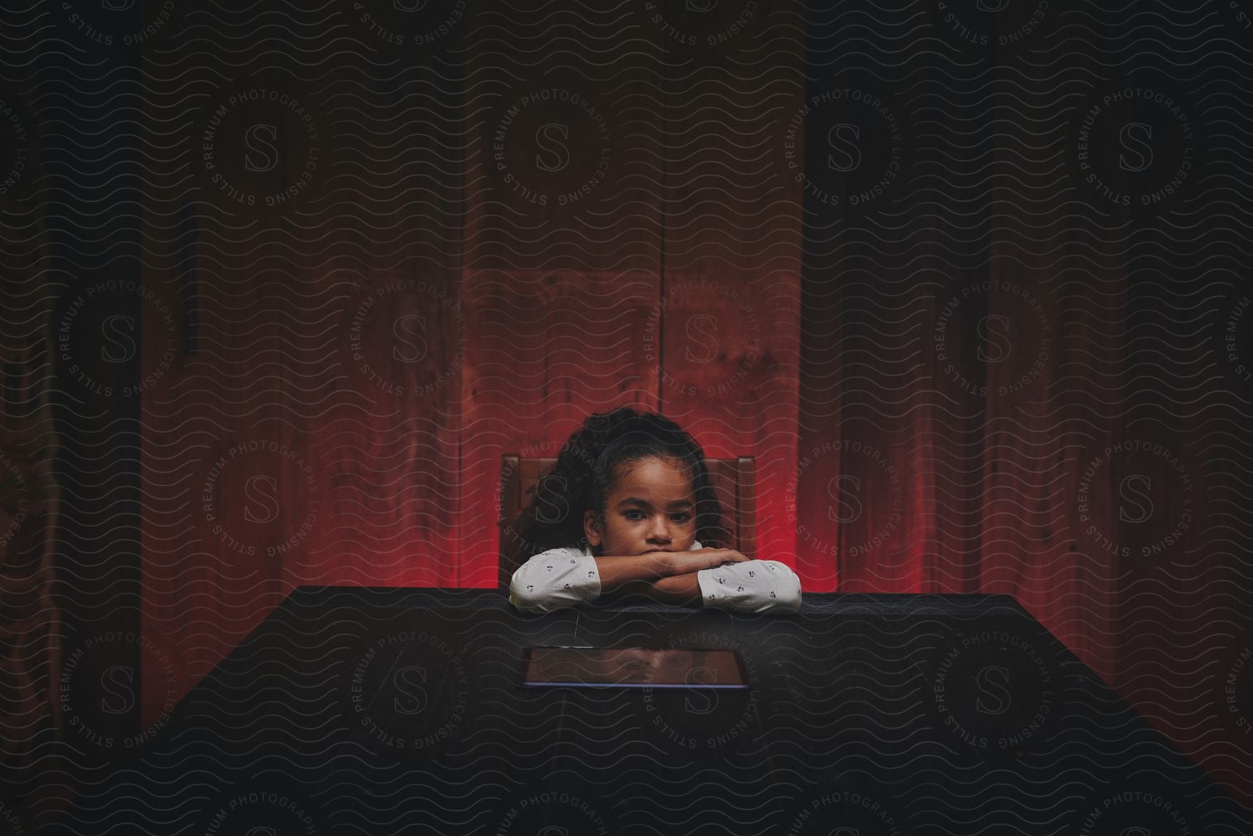 A young girl is sitting at a table with her arms folded and her head down as she looks at a tablet computer in front of her