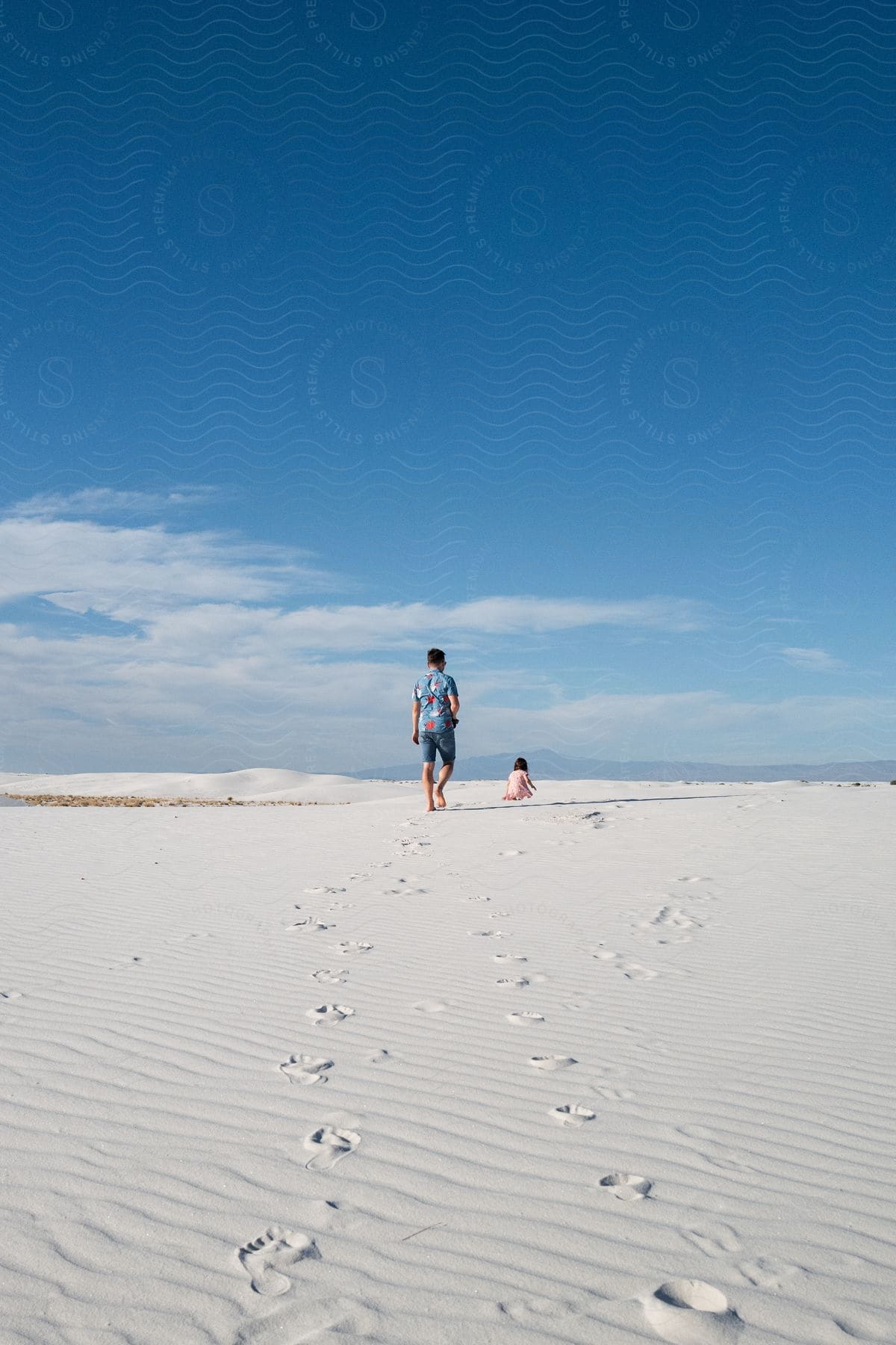 A man and a young girl walking on white sand under a clear blue sky, leaving a trail of footprints behind them.