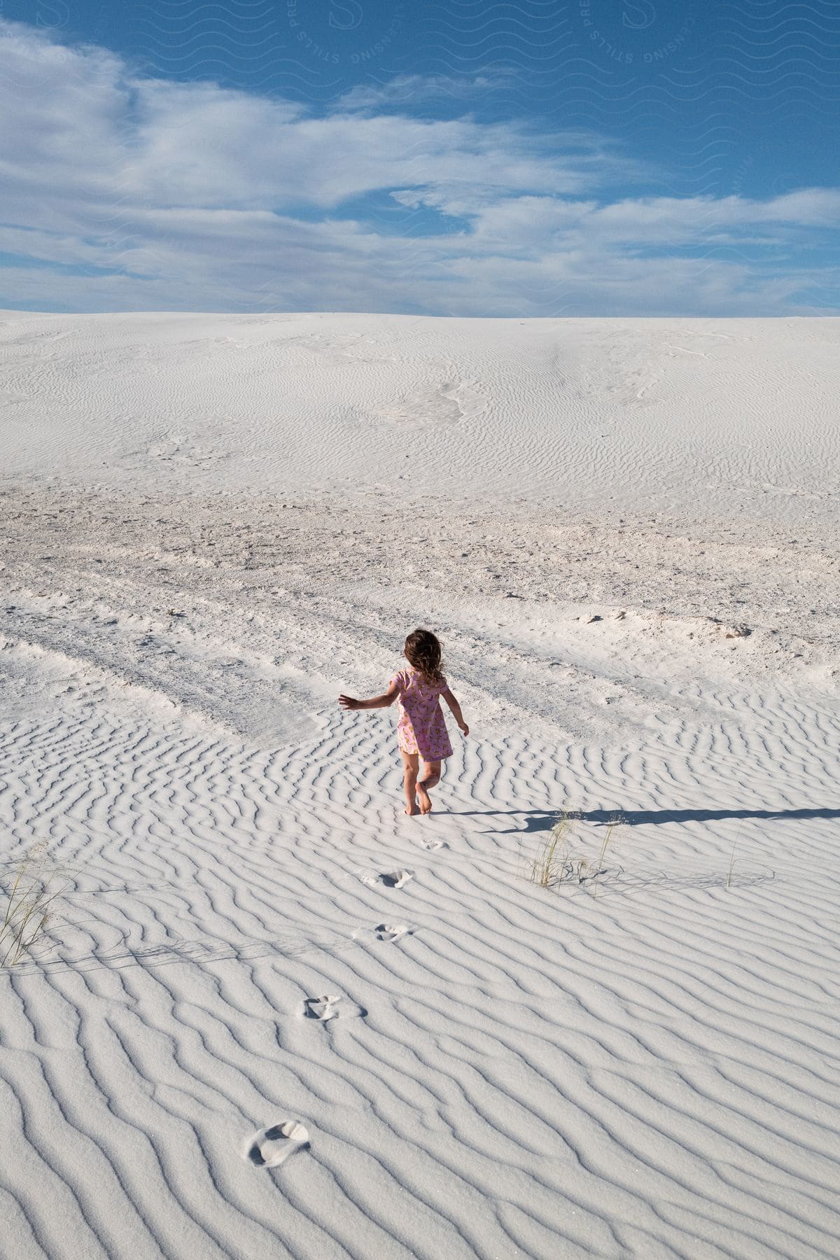 Little girl wearing a dress walks barefoot across white sand dunes under a cloudy sky