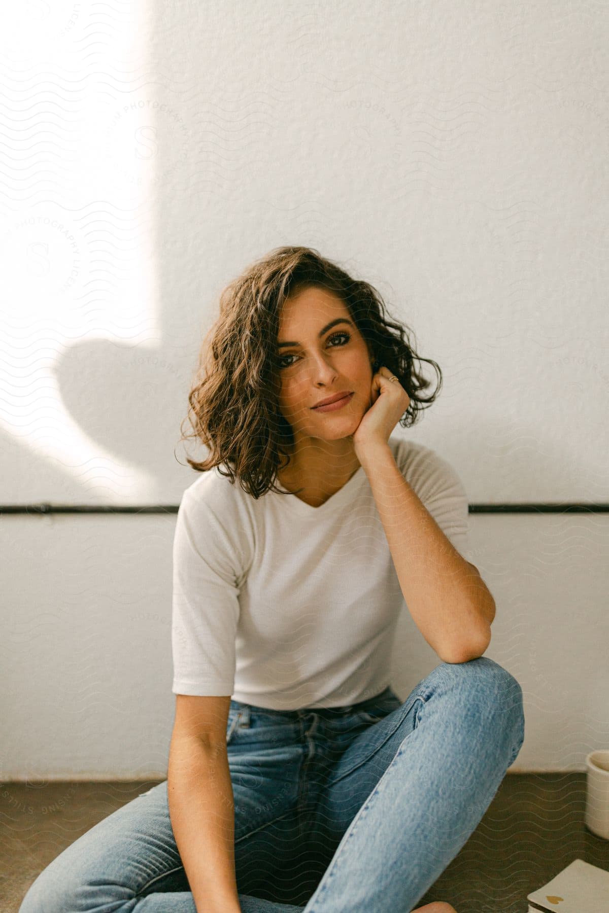 Woman with curly hair a white t shirt and jeans sits on the ground smiling at camera