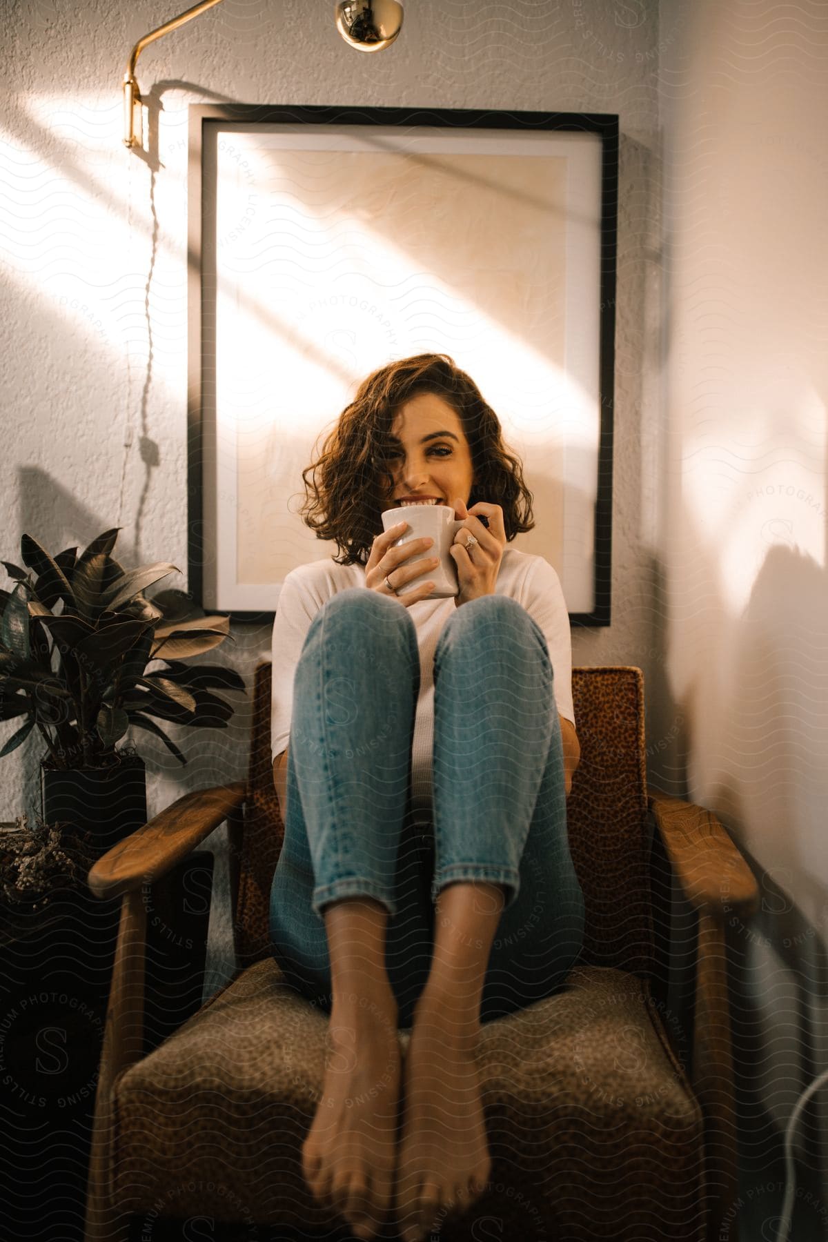 A brown haired woman in jeans sitting with a coffee mug in her hands and her knees pulled up to her chest.