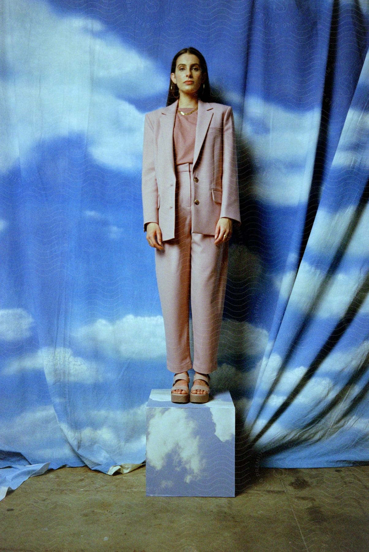 A woman with long dark hair wearing a pant suit is standing on a block in front of a sheet painted like the cloudy sky