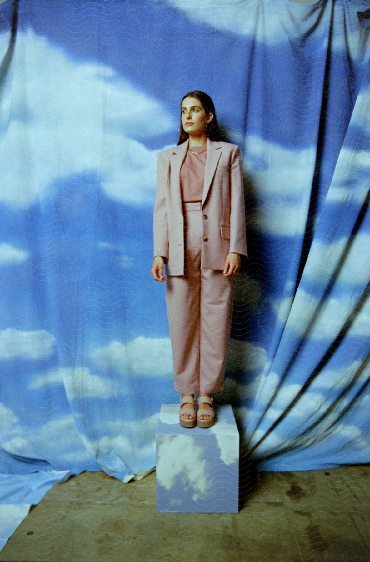 A woman with long dark hair wearing a pant suit is standing on a block in front of a sheet painted like the cloudy sky