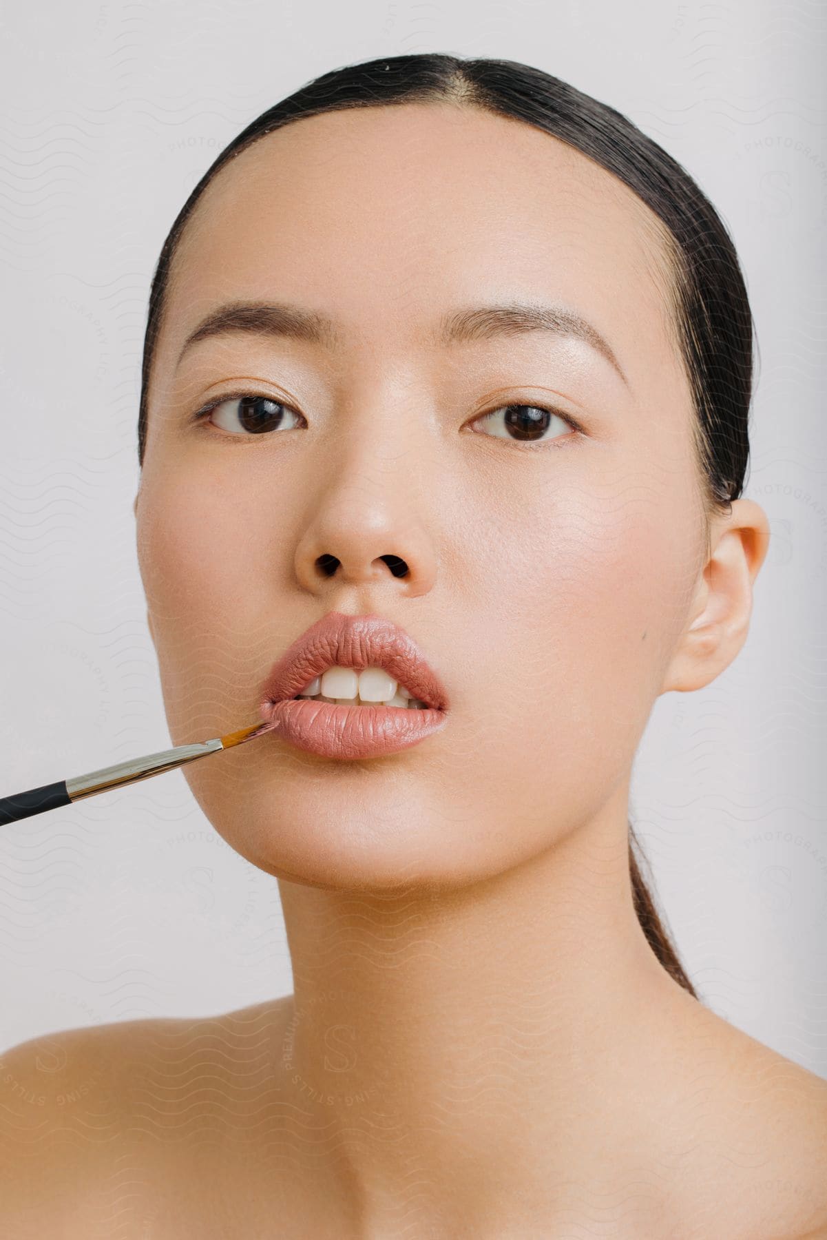 Portrait of a young Asian woman with a makeup brush on her lips.