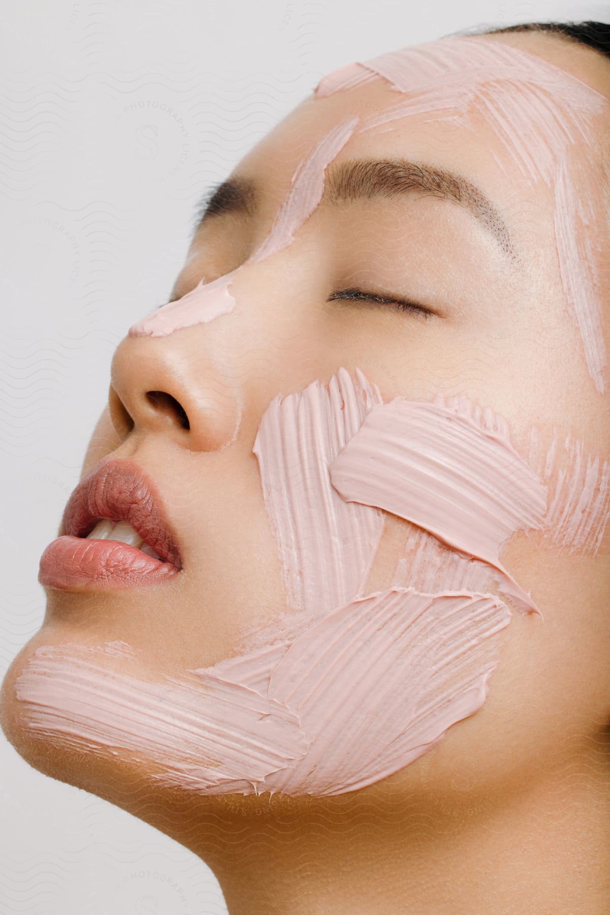 A young woman with stripes of skin cream on her face tilts her head back with her eyes closed