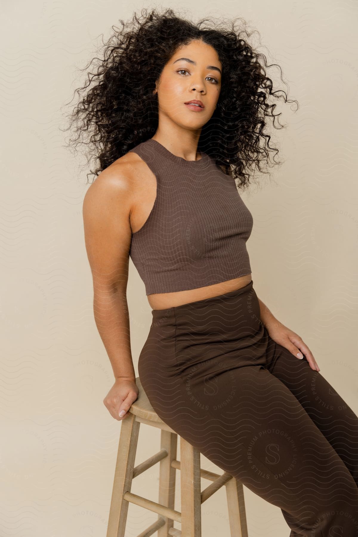 A young woman with dark, curly hair poses while sitting on a wooden bar stool.