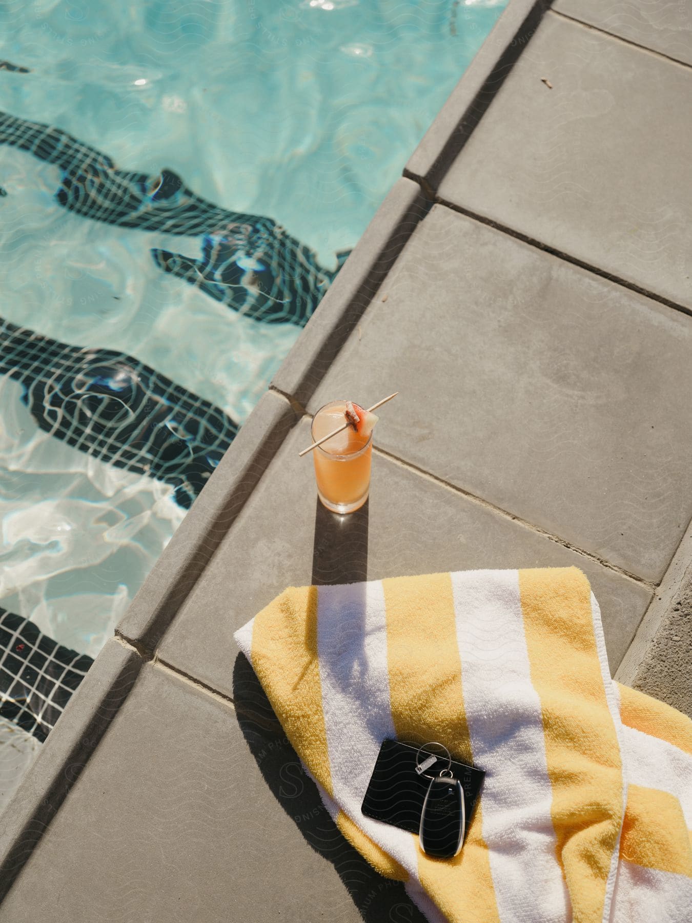 Car keys and a wallet on a towel near a glass of drink by the swimming pool.