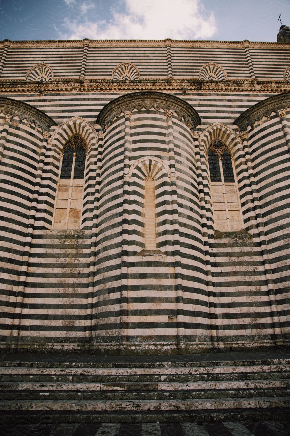 Striped lateral of a cathedral with arched windows and weathered steps, set against a blue sky with clouds.