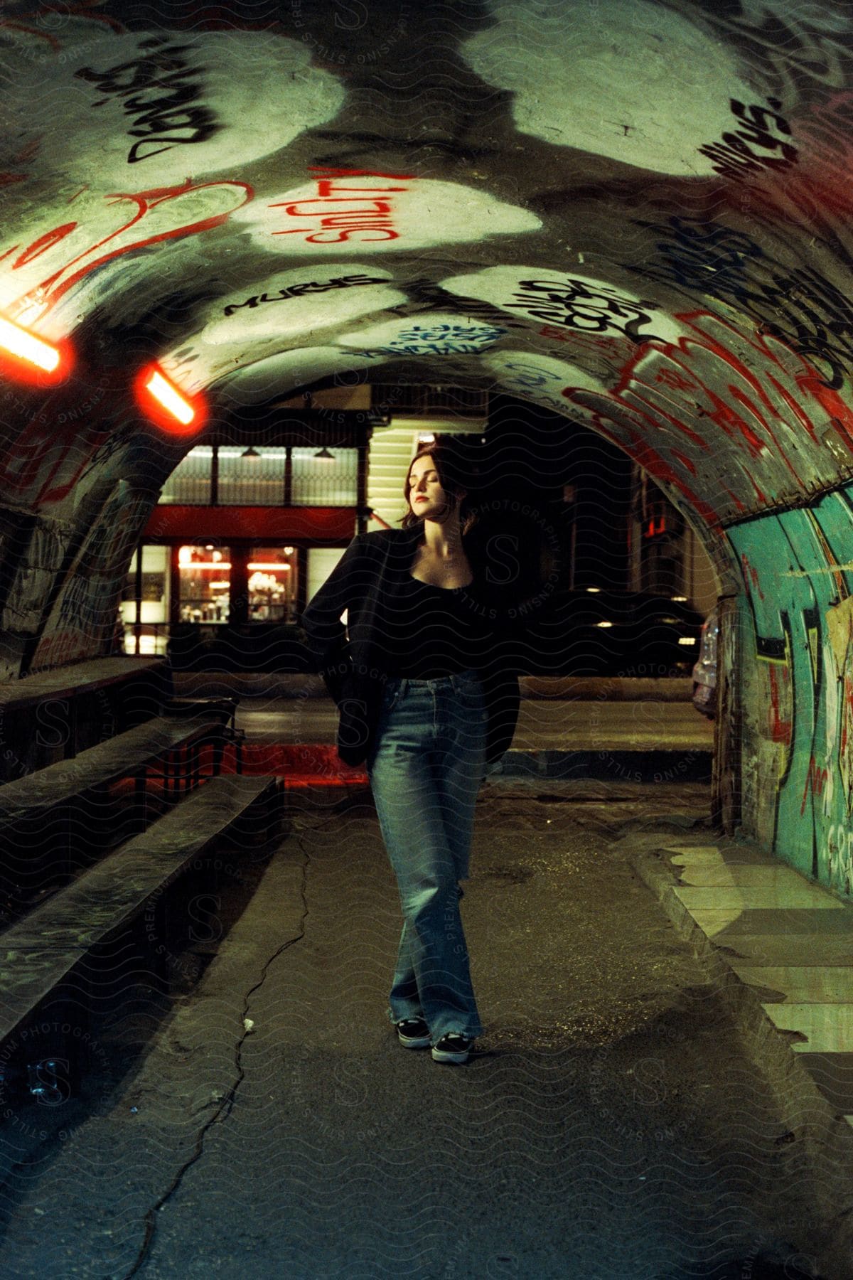 A Woman Wearing A Black Top, Jacket, And Jeans Stands In A Tunnel Adorned With Graffiti