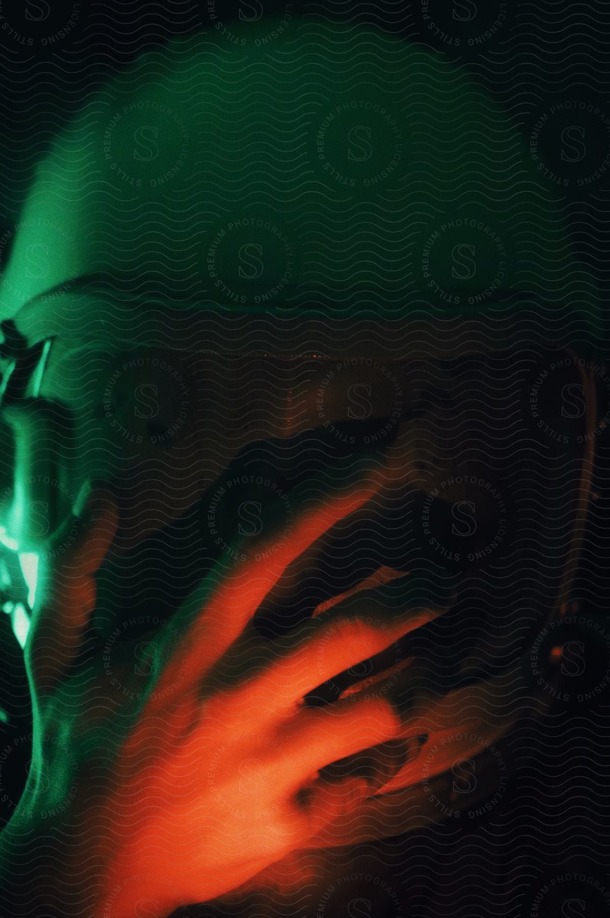 Red light shines on a mans hand in front of the VR helmet he is wearing in the darkness as green light shines in the distance