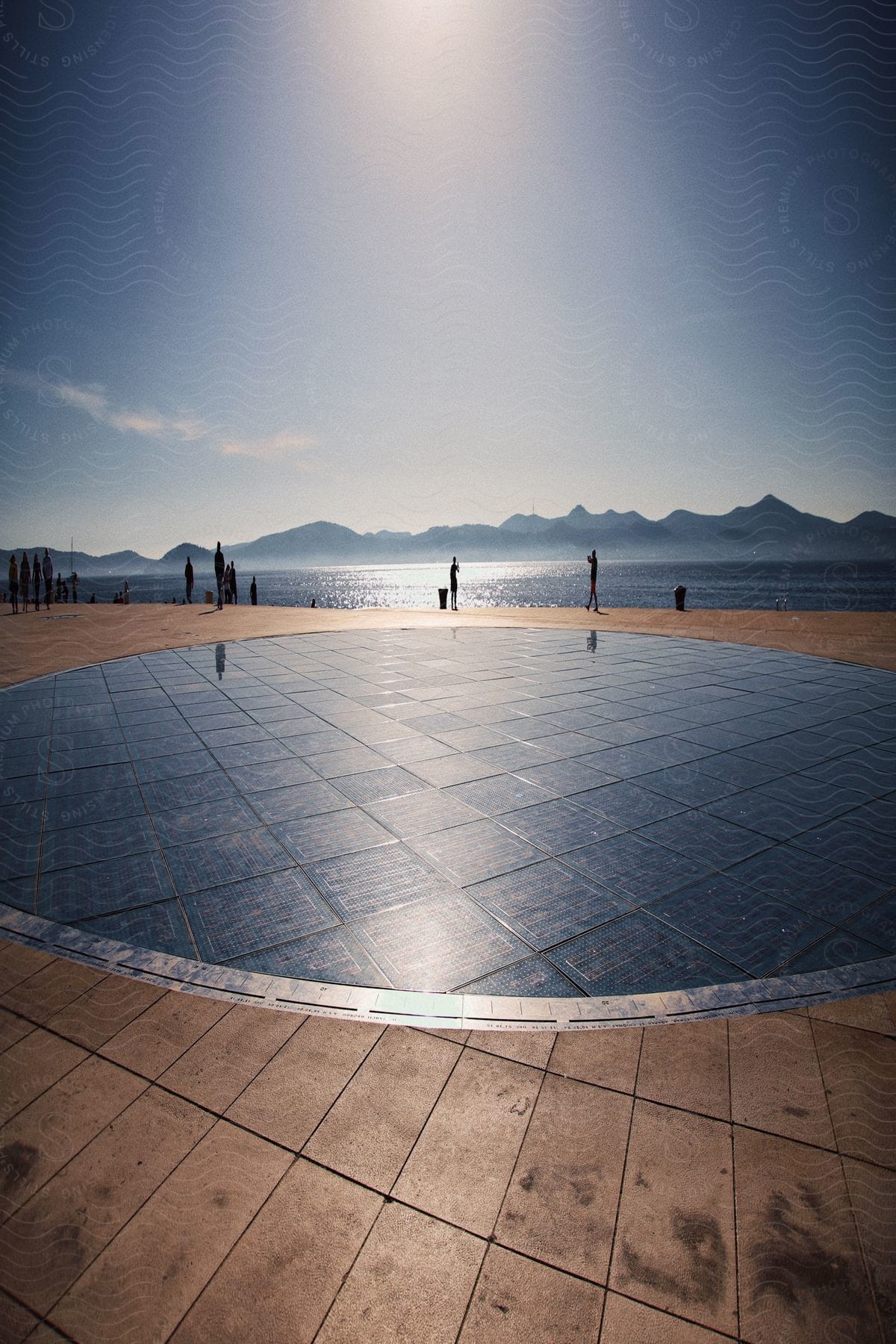 Silhouettes of people walking near the Monument to the Sun along the coast of the Zadar peninsula in Croatia with mountains in the distance