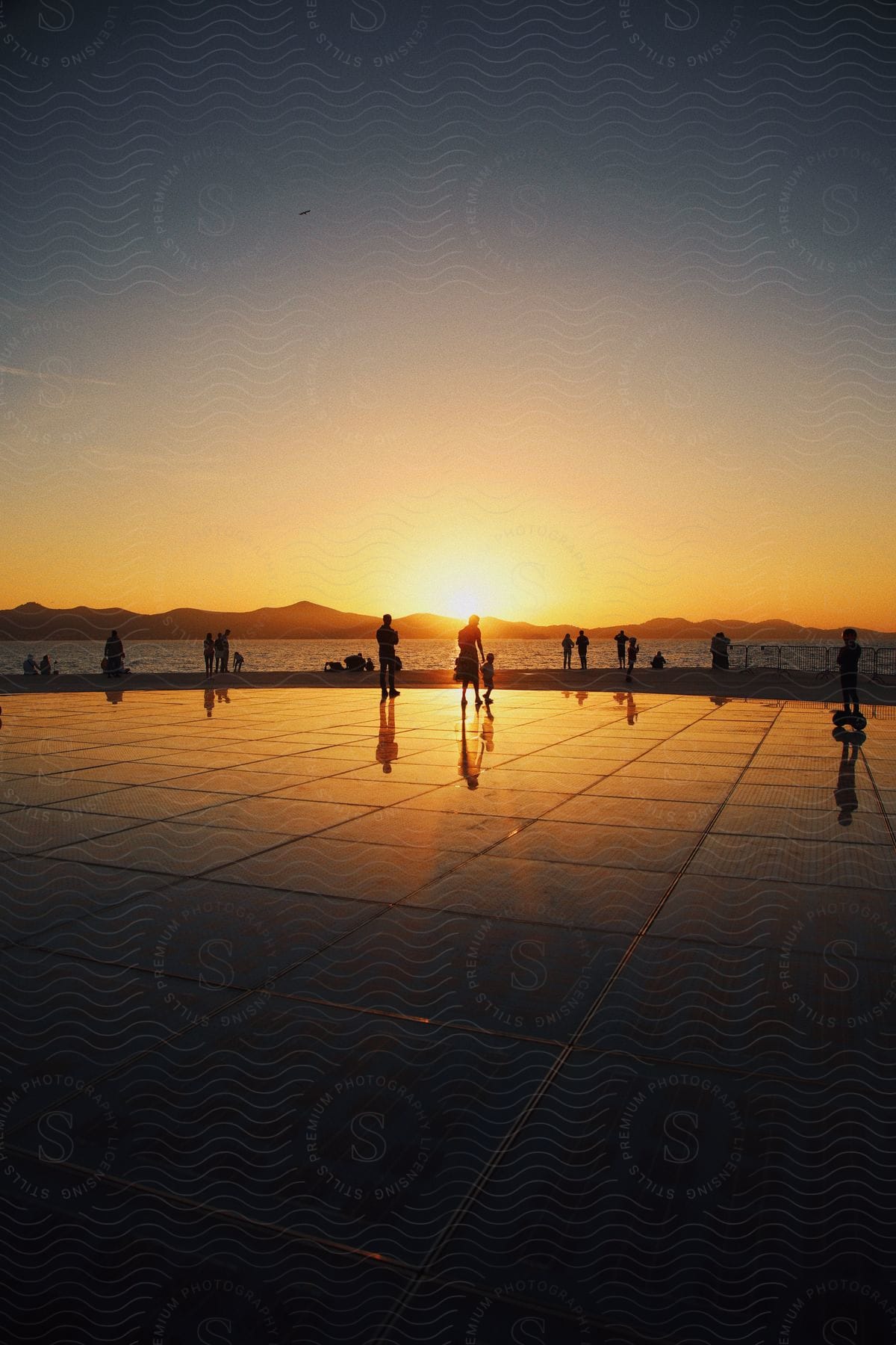 Sunset on a reflective promenade with silhouettes of people against a backdrop of distant ocean, mountains and a golden sky.