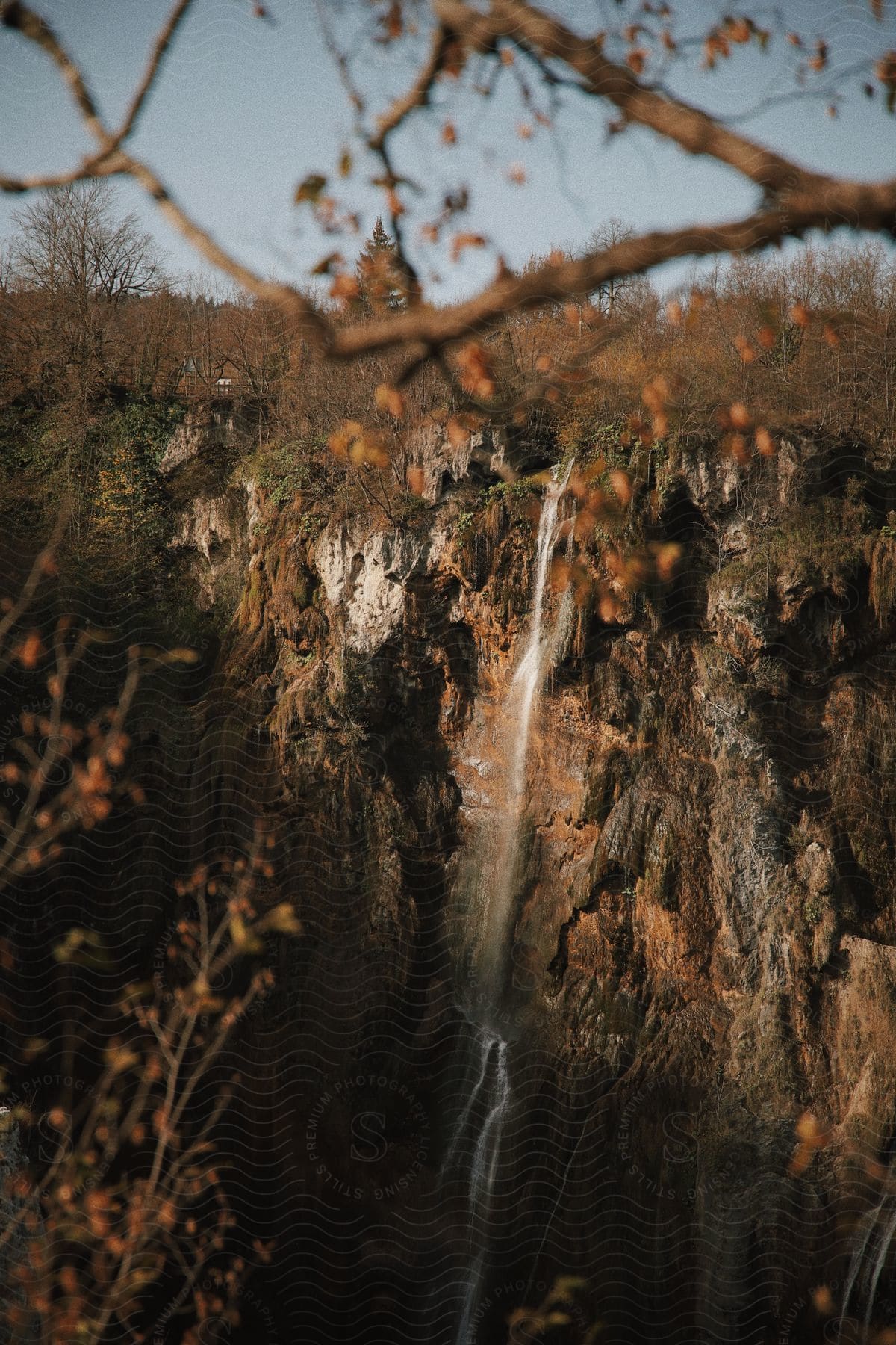 A small waterfall spills over a steep cliff with a deciduous forest in the background during late fall.