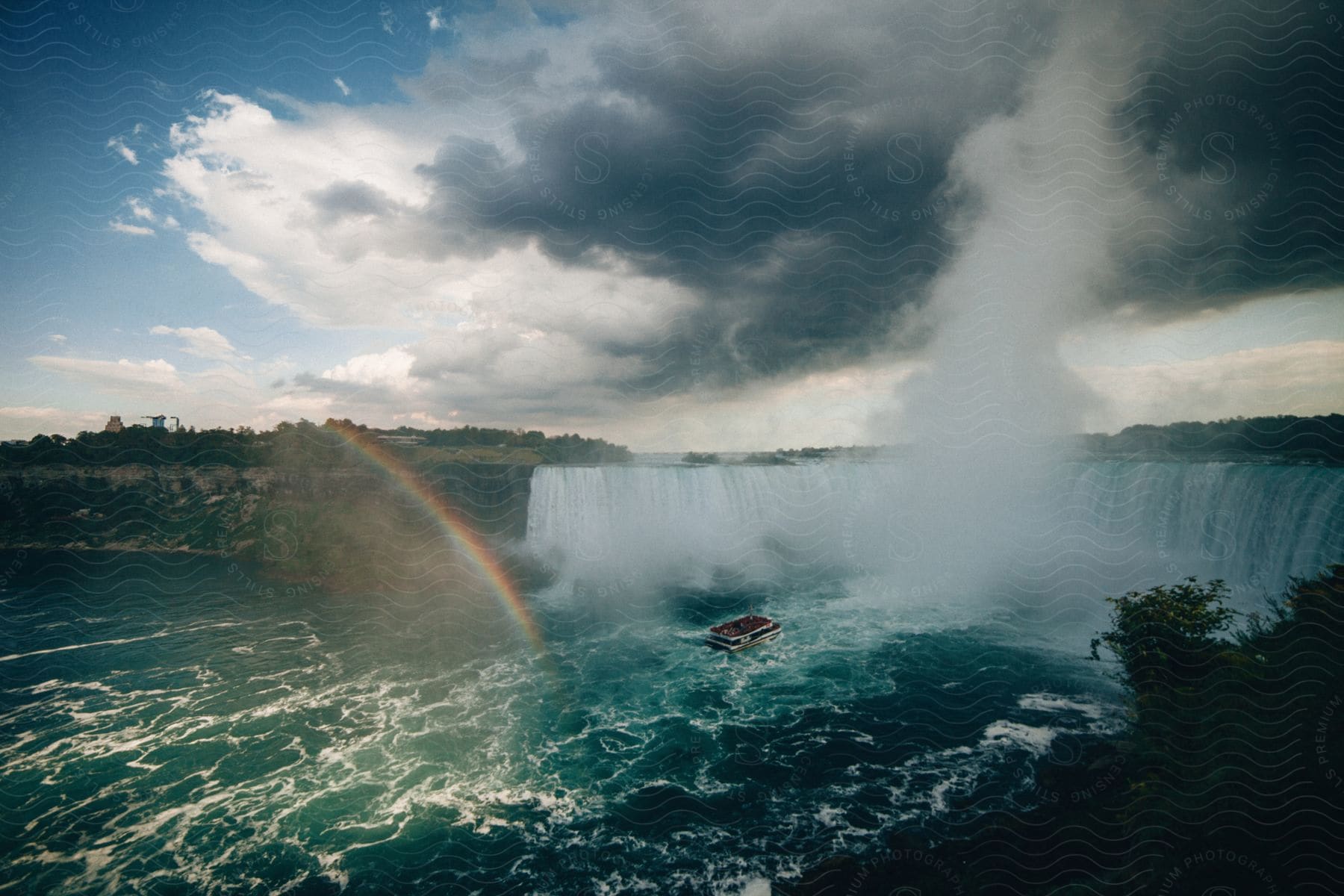 Boat floats between large waterfall and rainbow under  heavy clouds.