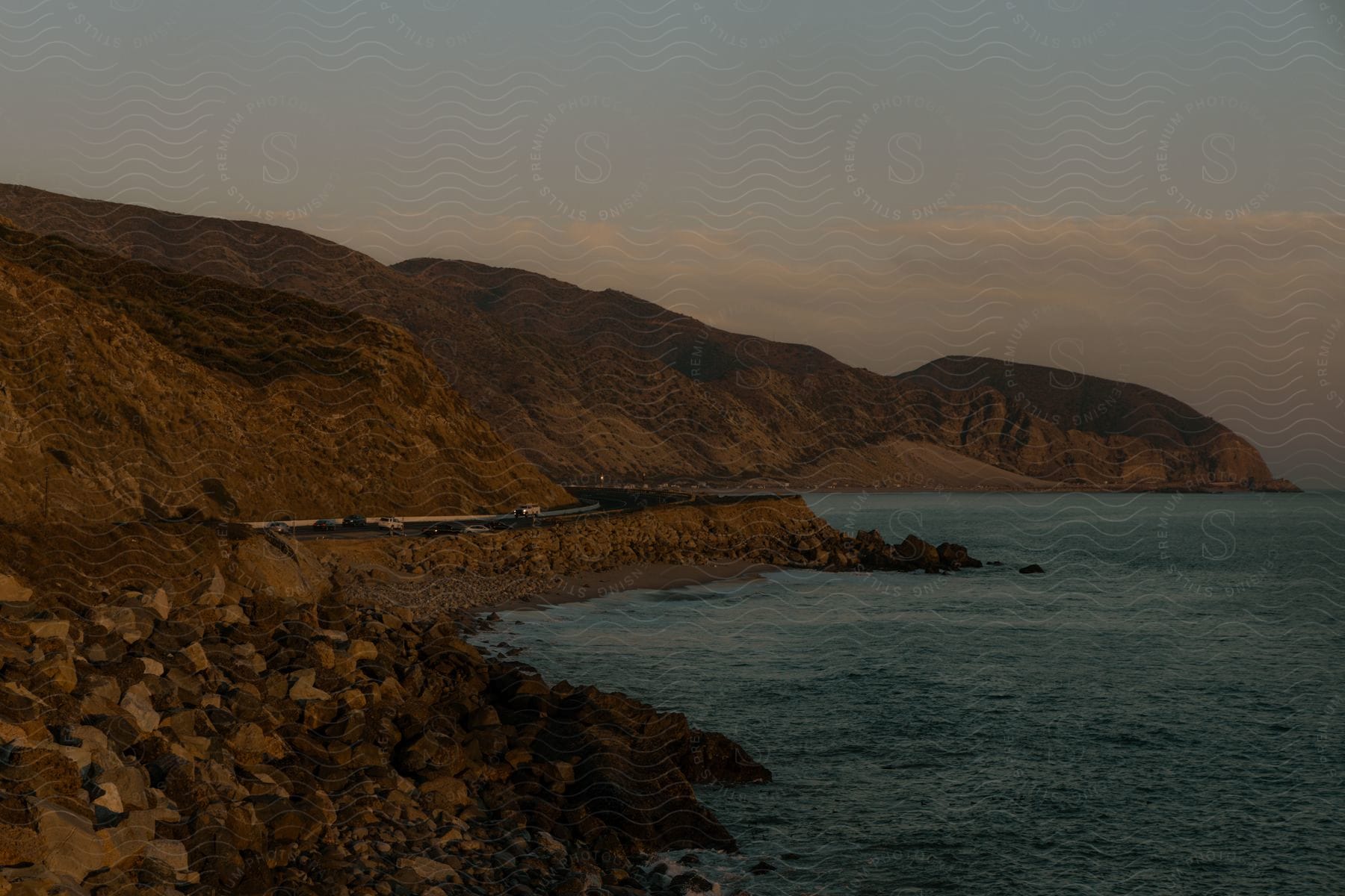 Coastal road winding along the base of rolling hills right next to the seashore, during twilight with soft light.