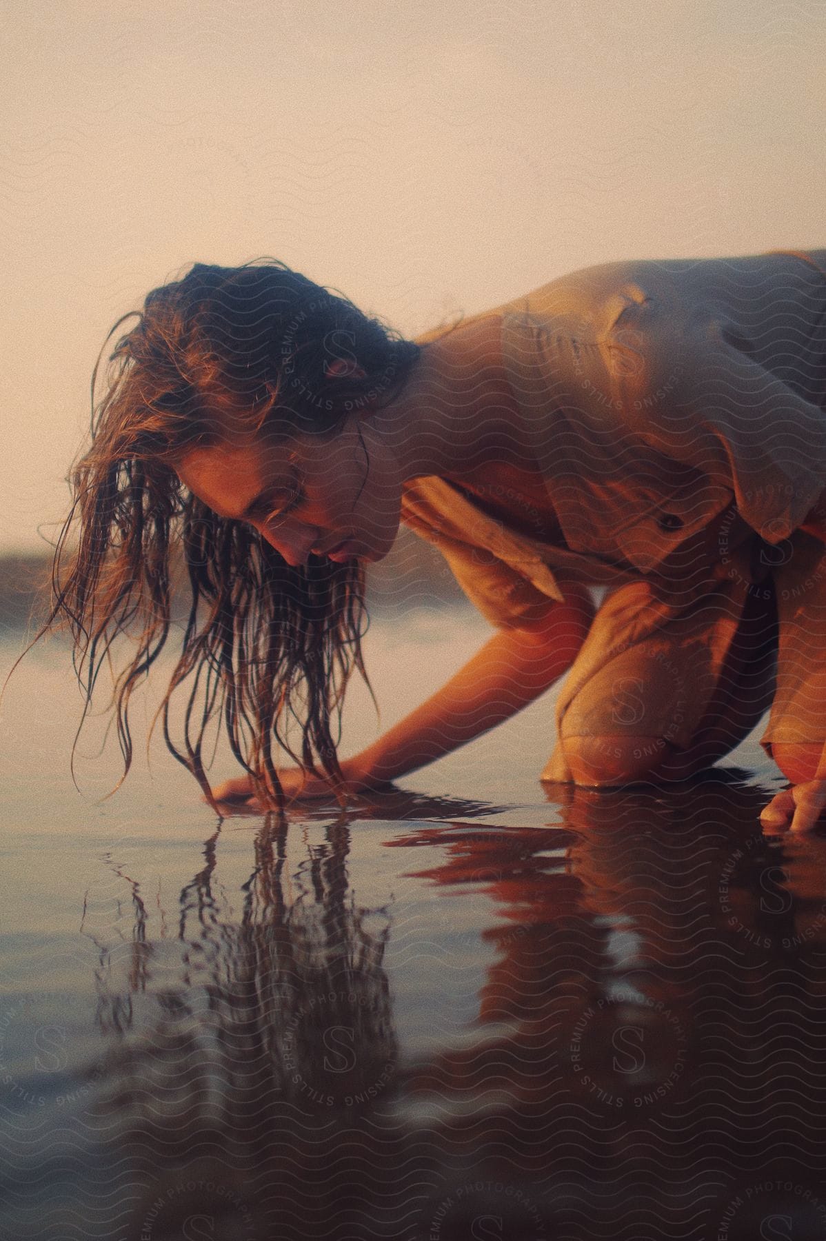 A woman with long hair kneeling on a beach in shallow water while looking down.