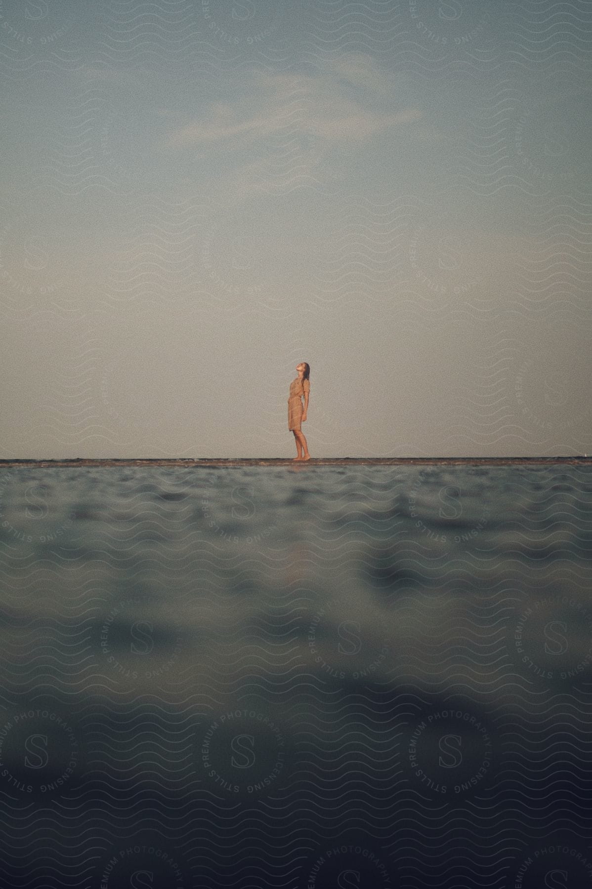 A woman wearing a beige dress is standing on the beach near the water and looking up at the sky.