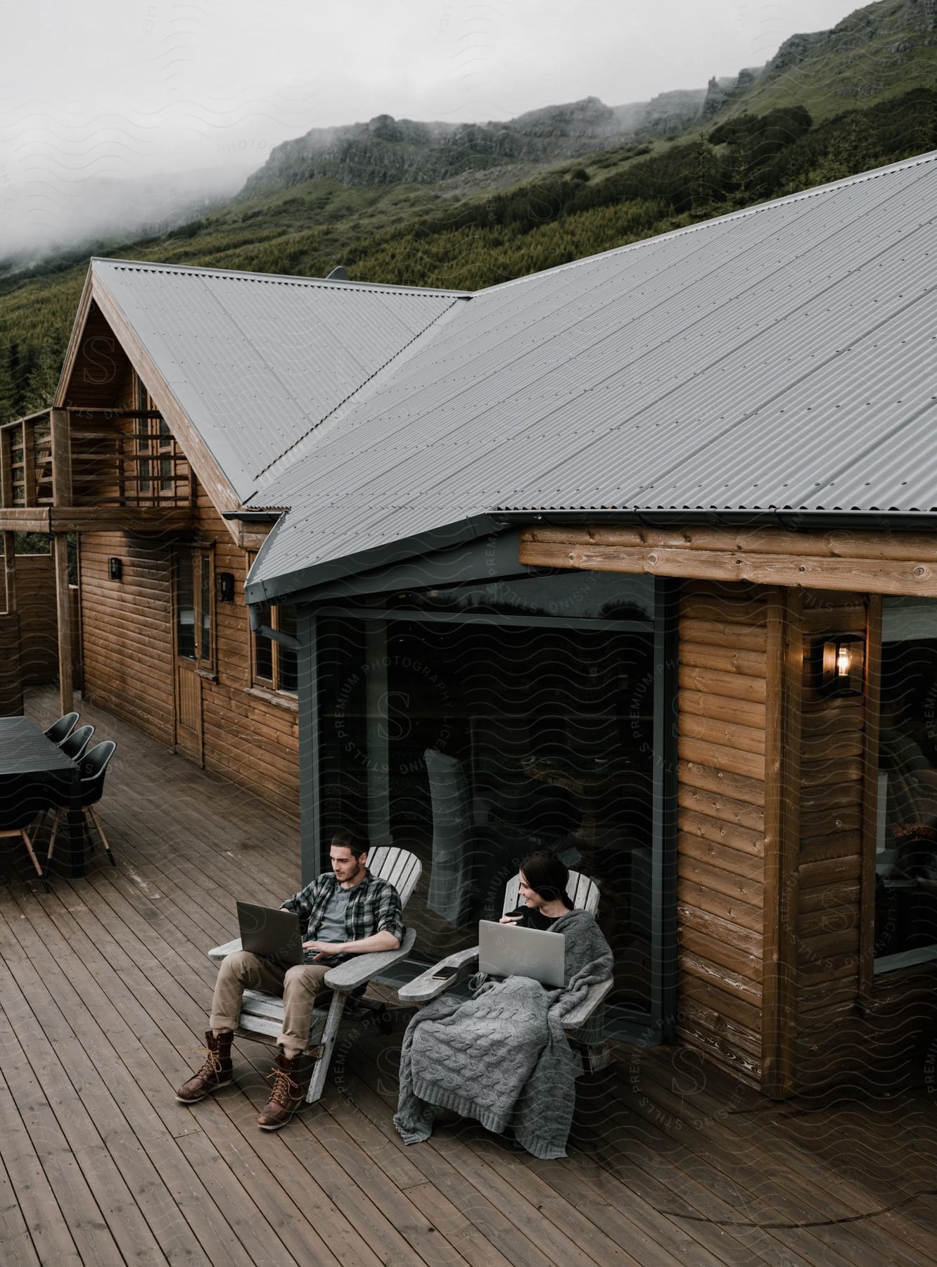 A couple sitting on the deck of a log cabin both holding laptops with fog on the mountain ridge behind them