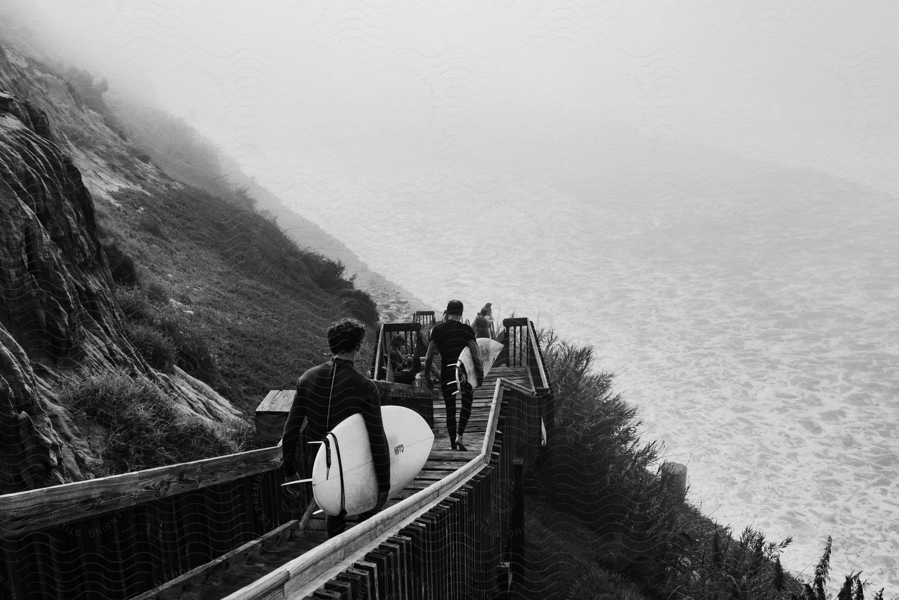 Two surfers, clad in wetsuits and carrying surfboards, walk down a wooden staircase to the beach.