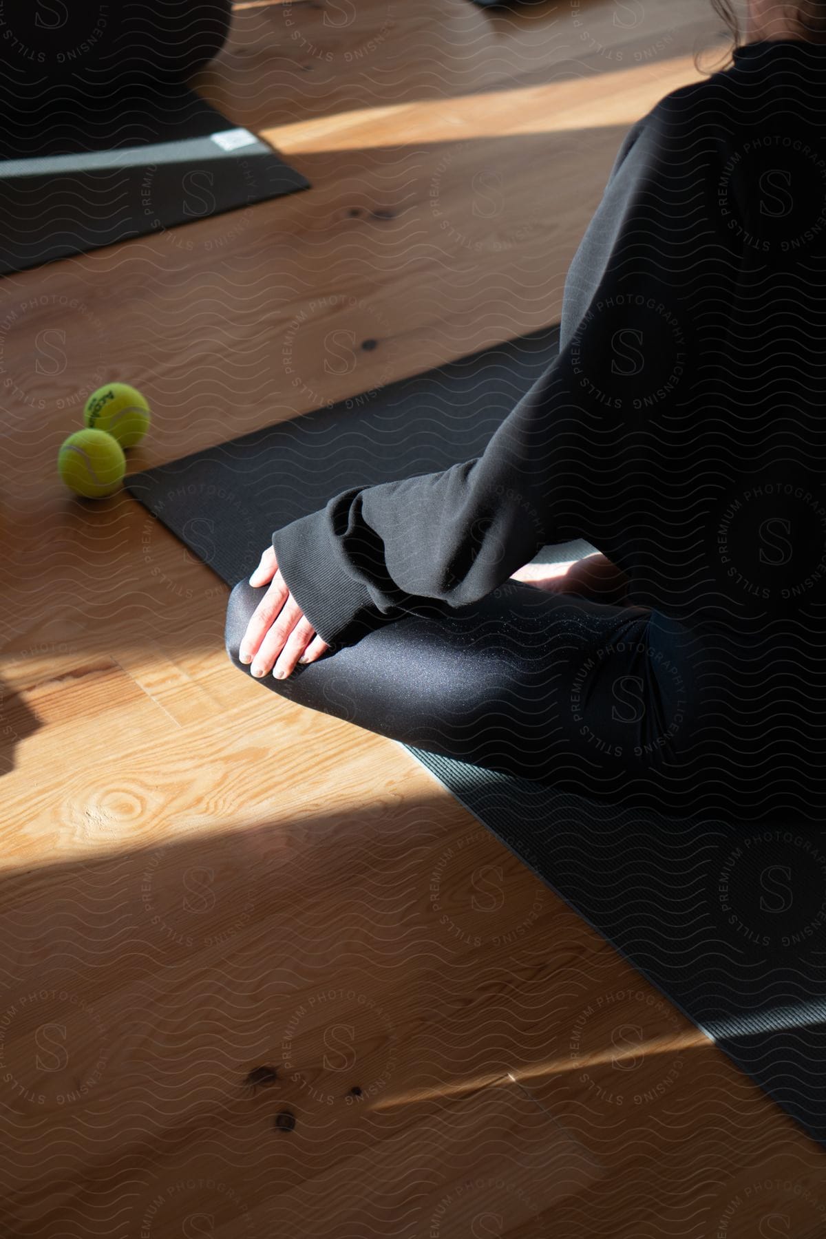 A person seated on a yoga mat on a meditative pose, hands resting on their knees, sunlight streams in, with two tennis balls lying nearby on the wooden floor.
