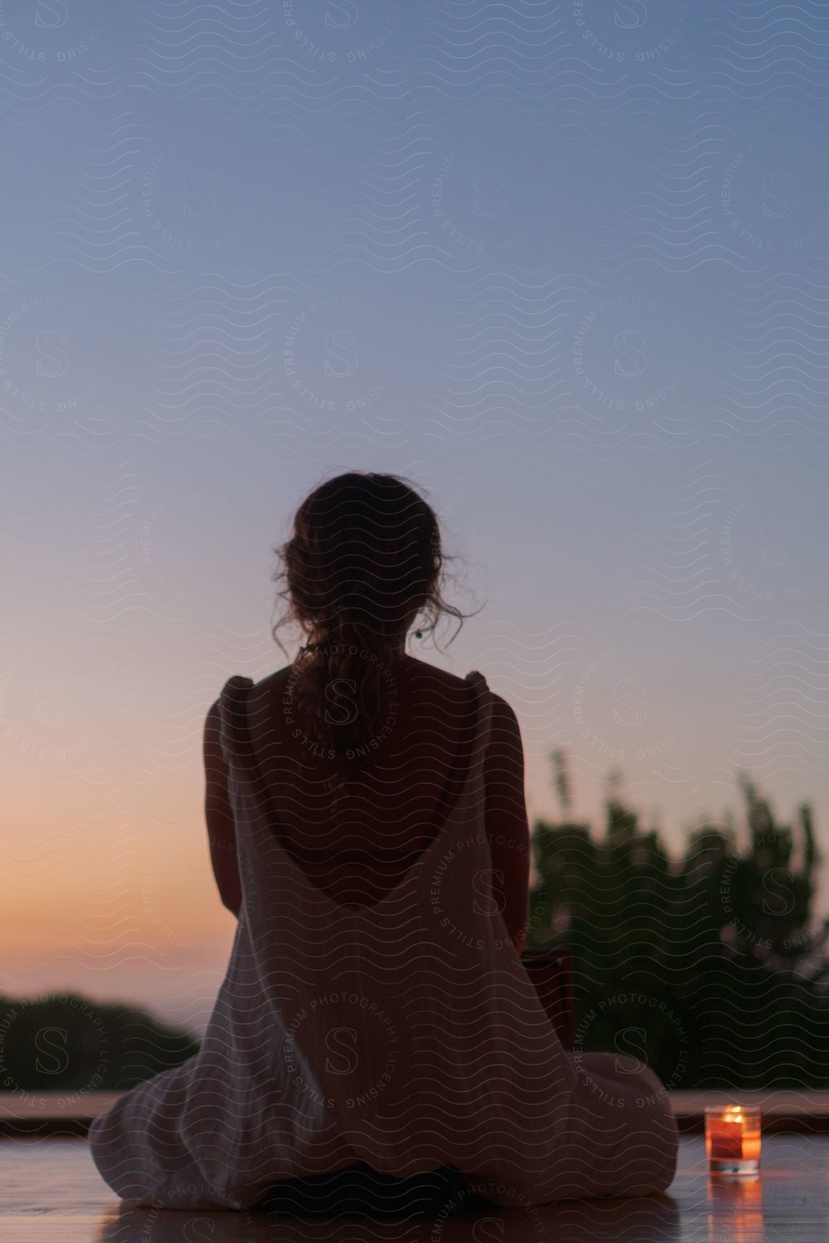 Woman sitting in a yoga position with her back to the viewer, a candle beside her, and a reddish dawn sky.