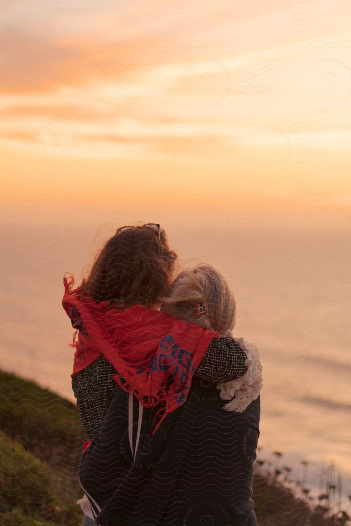 Mother and daughter hugging each other with their backs to the camera and facing the sea with a reddish dawn sky.