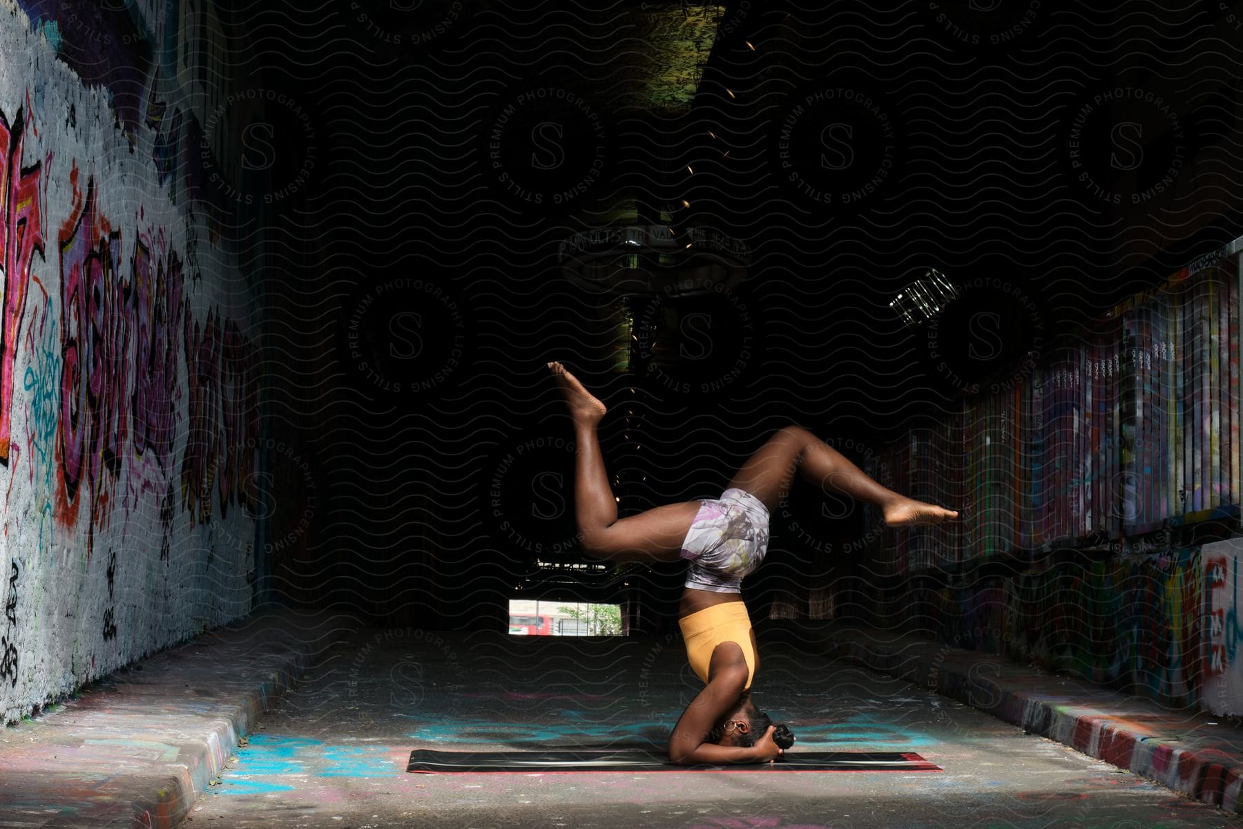 A black woman in a forearm hollow back yoga pose on a mat in a tunnel with graffiti on the walls