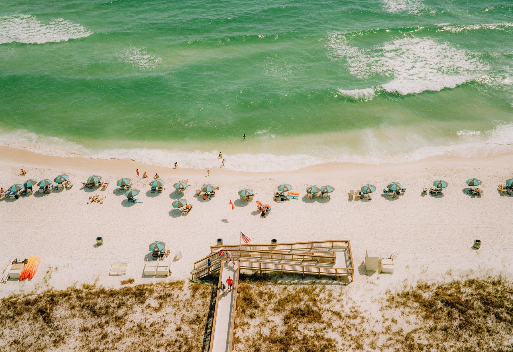 Aerial view of beachgoers on white sandy shores with turquoise waters, umbrellas, and a wooden walkway providing access to the beach.