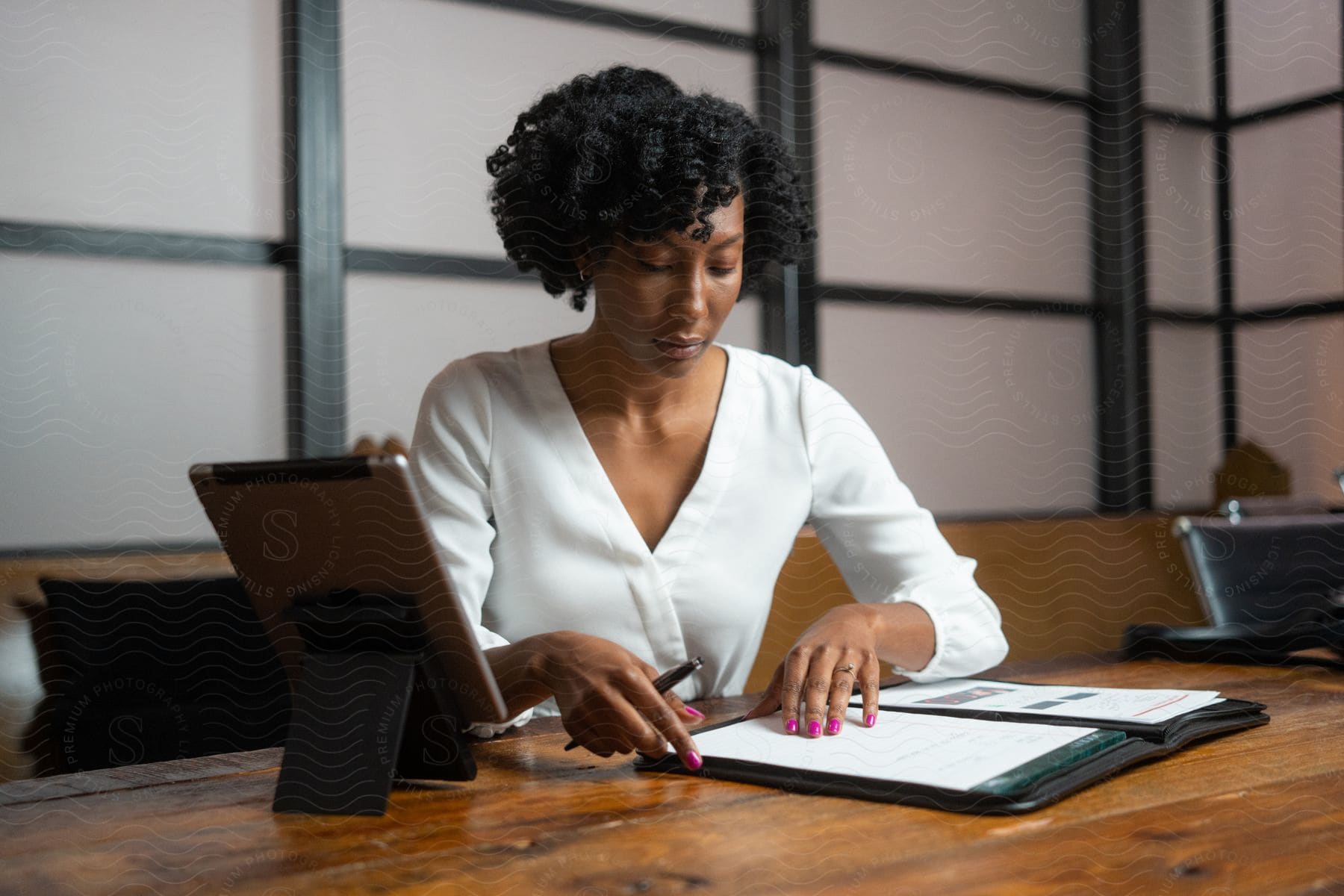 A businesswoman with curly hair and a white shirt sits at a desk next to a tablet in the office.