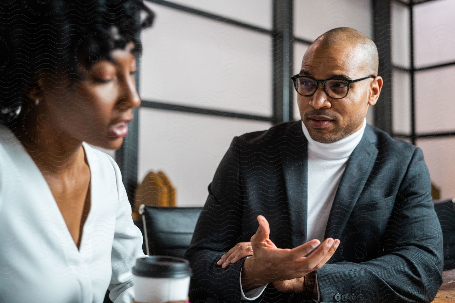 Businessman with glasses white turtleneck and a blazer jacket talks to businesswoman wearing a white shirt in the office