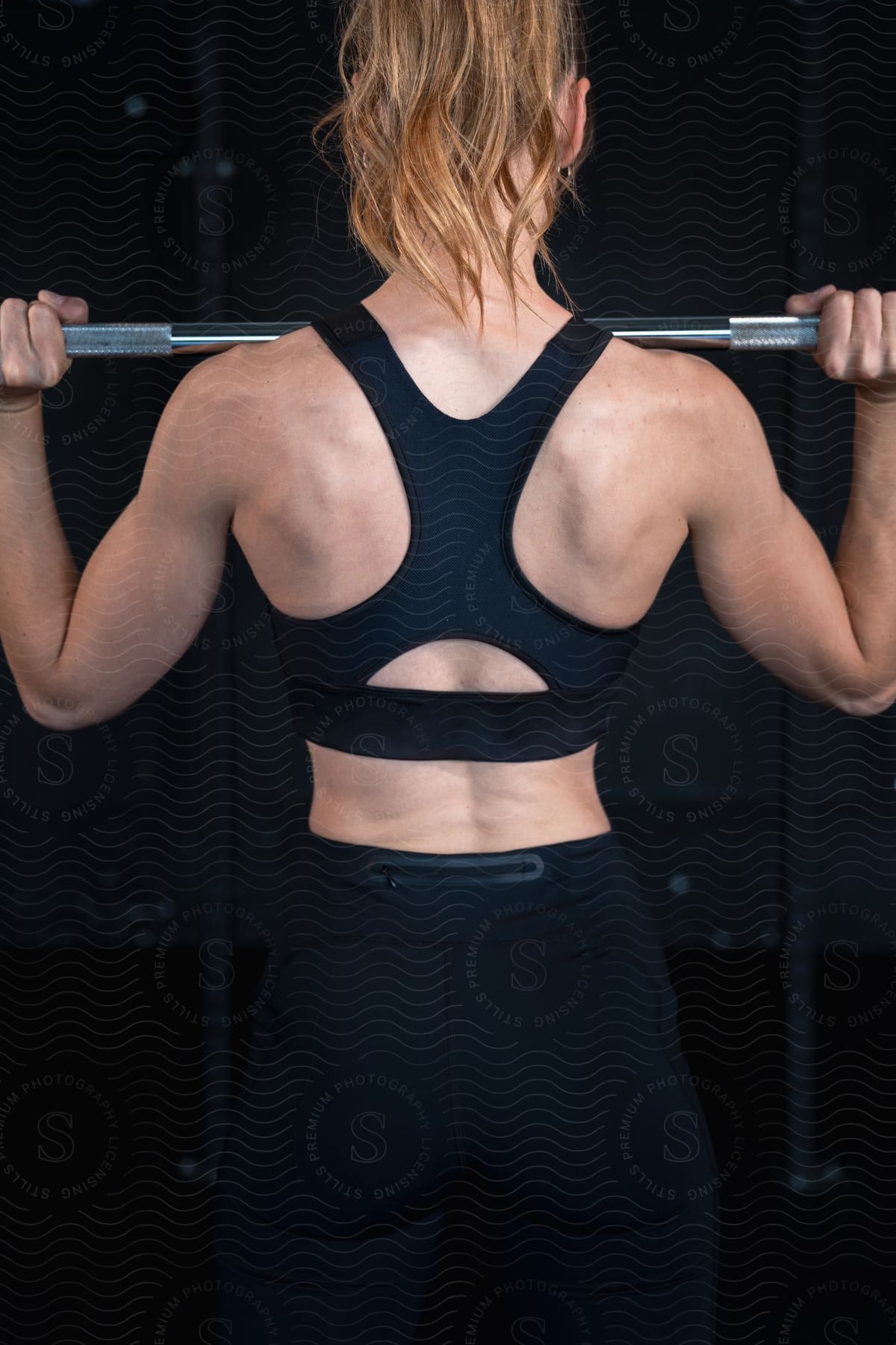 Rear view of a woman with a ponytail lifting a barbell, showcasing her muscular back and shoulders, dressed in athletic wear.