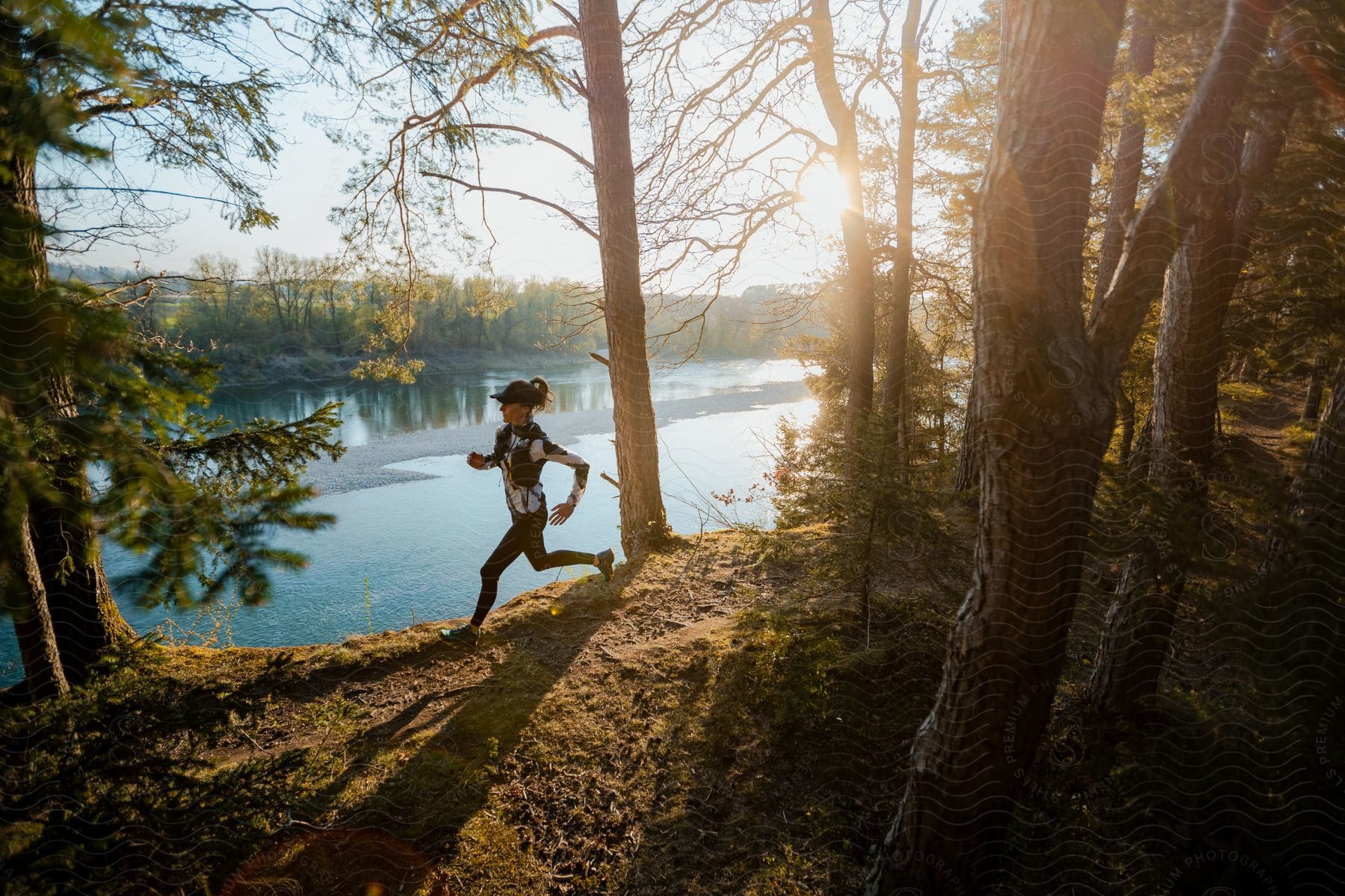 A woman with a cap, a white and black sweater, and black leggings runs in the forest near a lake, with the sun shining in the background.