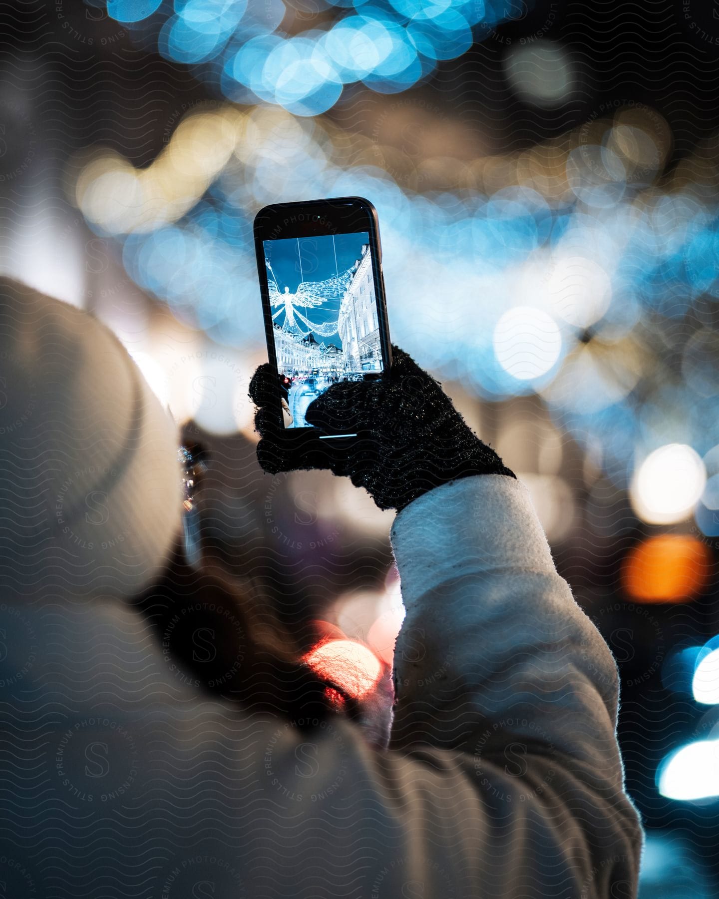 Stock photo of a woman wearing winter clothes holds up her cellphone to take a picture of holiday lights in the city