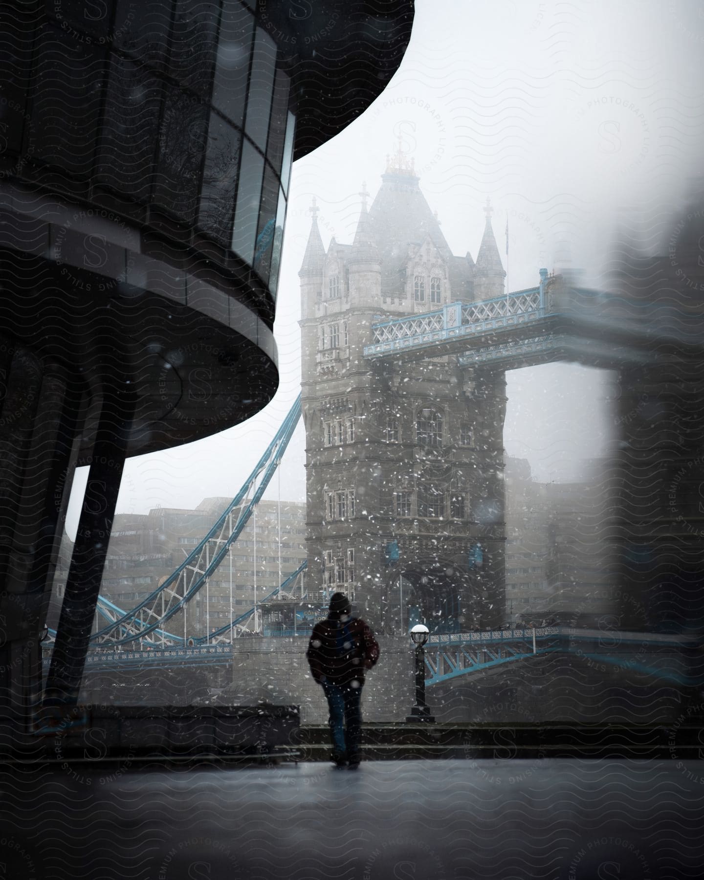 A man wearing a black hoodie is walking in front of the Tower Bridge, on a snowy, foggy day.