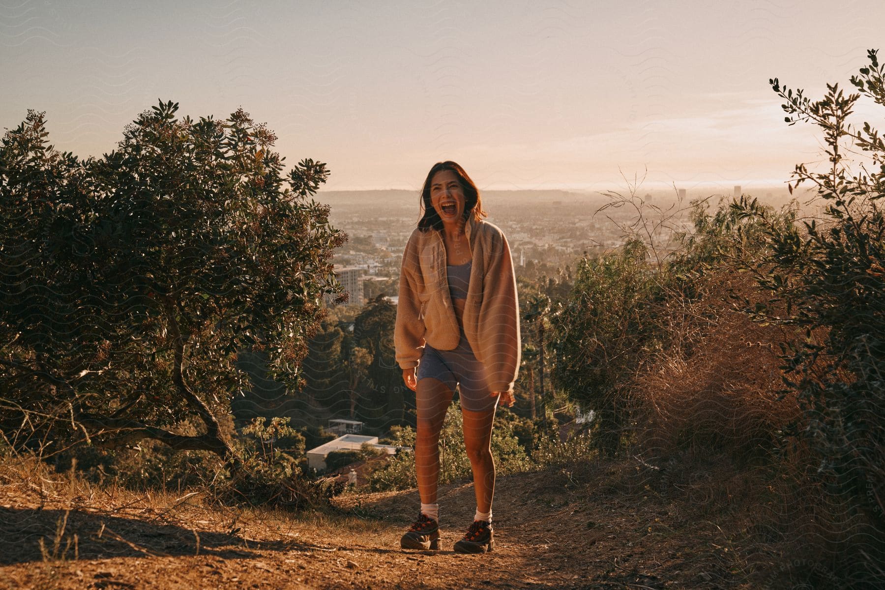 A woman wearing a white jacket, gray sports bra, and shorts walks uphill at sunset, with a city skyline in the background.