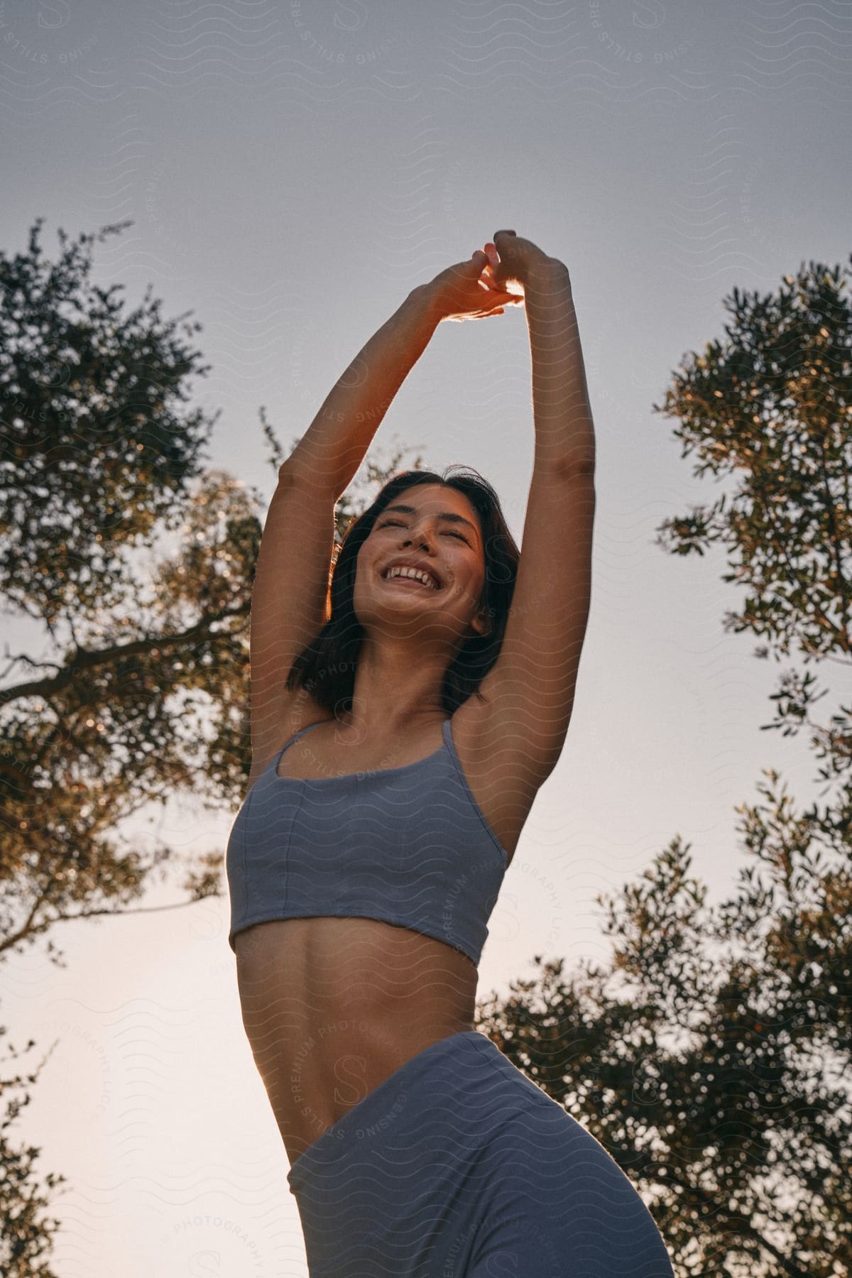 Smiling woman with arms raised in sportswear against an sky and tree silhouettes.