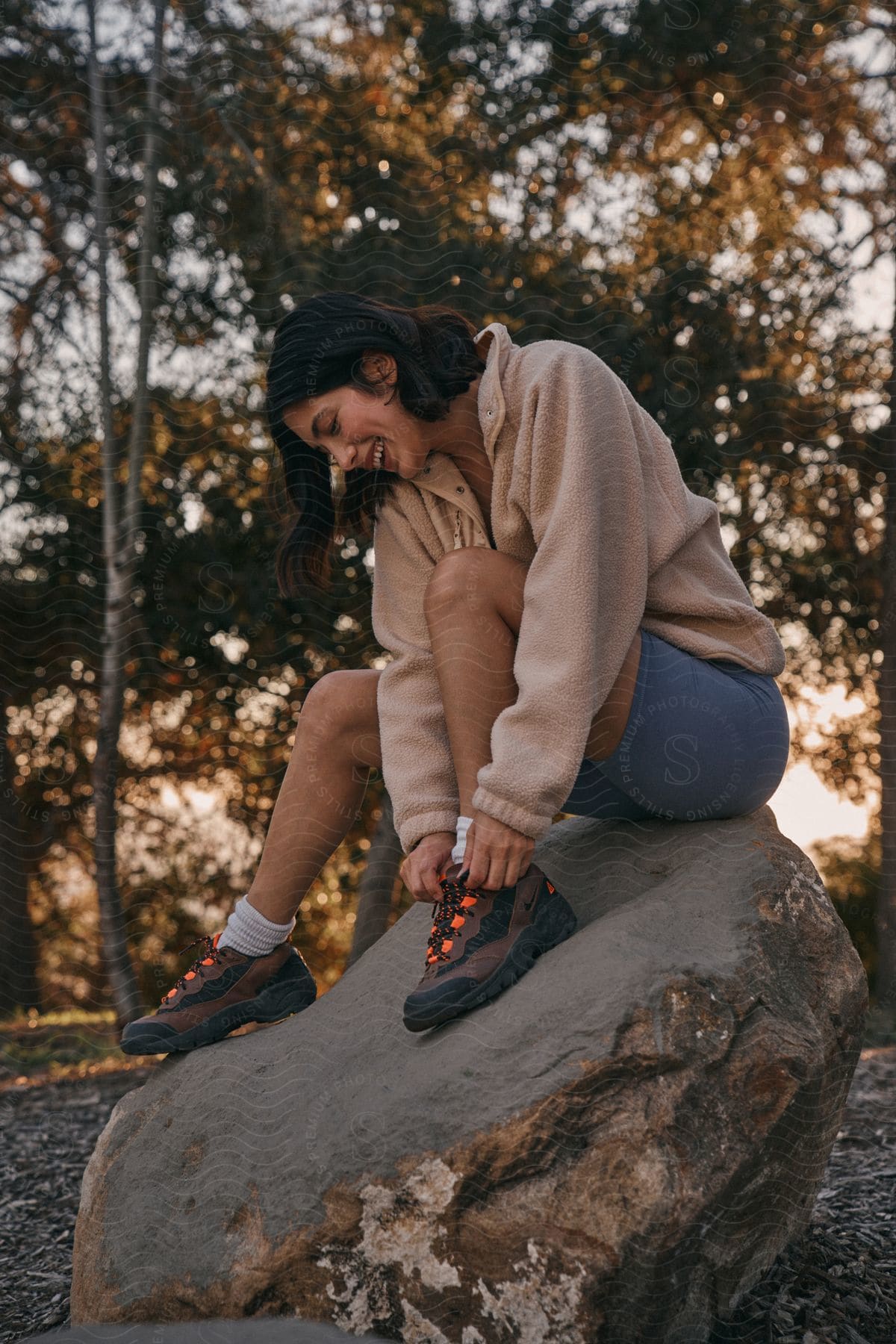Woman tying shoe laces on a rock outdoors with trees in the background at dusk.