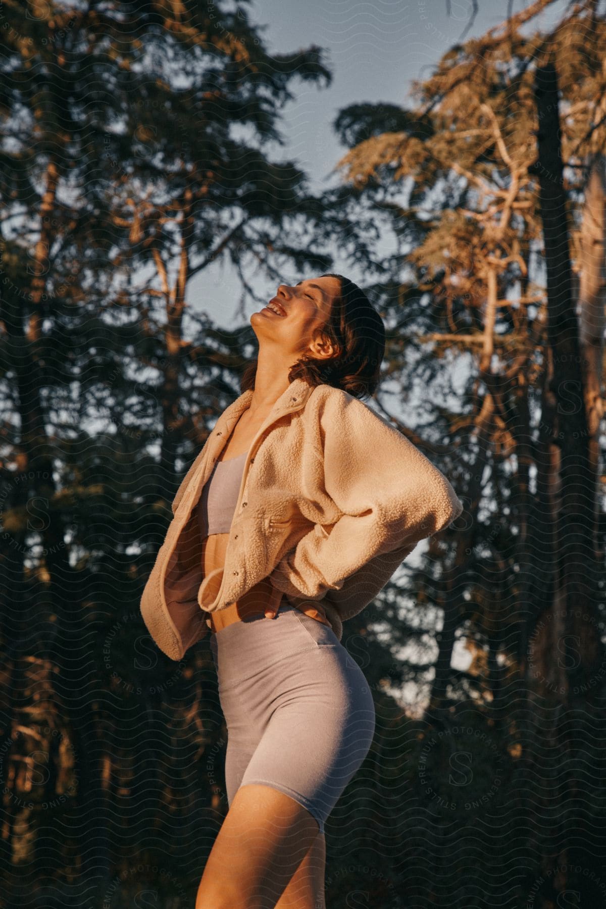 Woman modeling with hands on waist, looking upward with a smile, wearing a fitness outfit and a wool coat in a natural environment with trees.