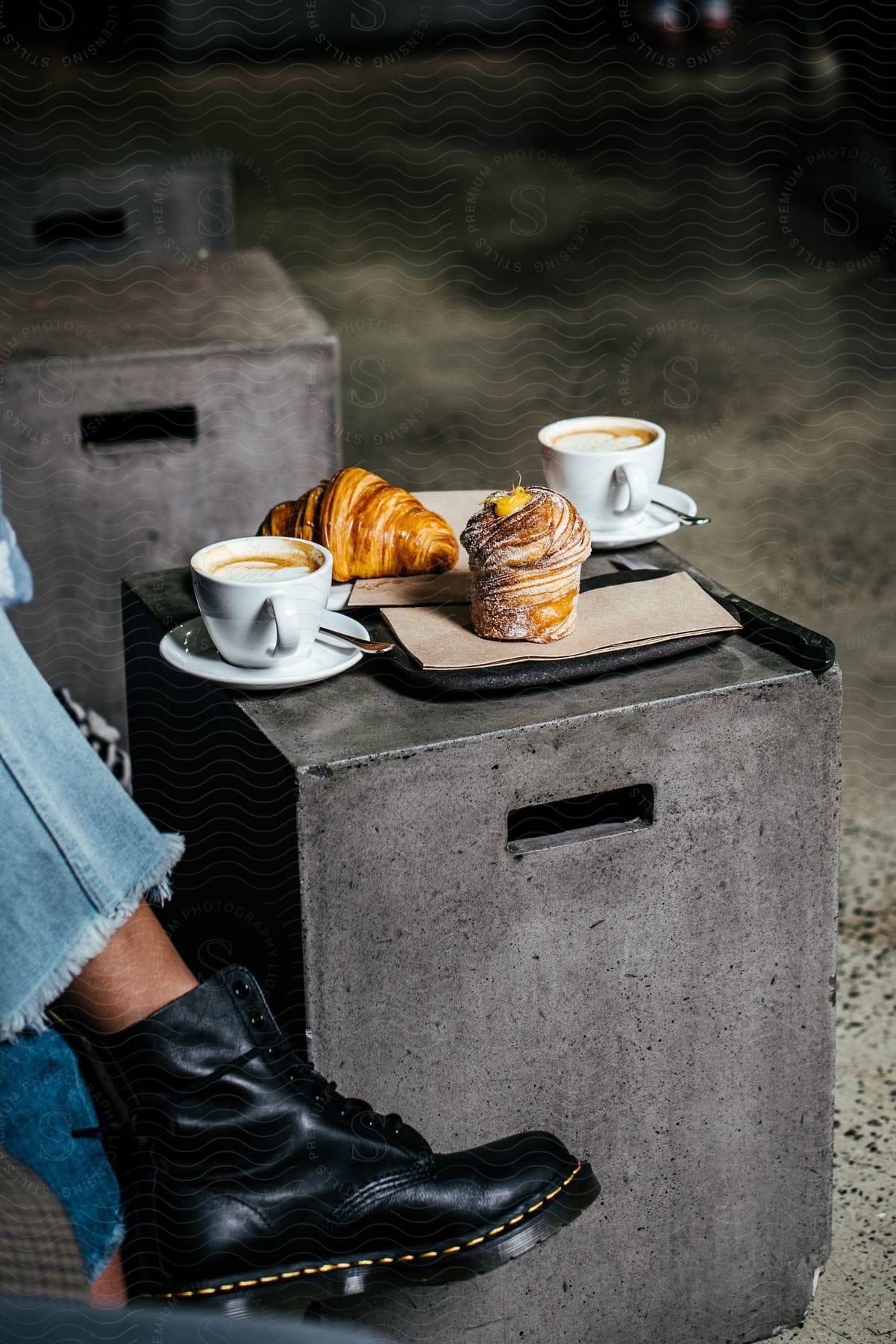 A mans foot is up near a tray with a croissant and muffin with two cups of coffee