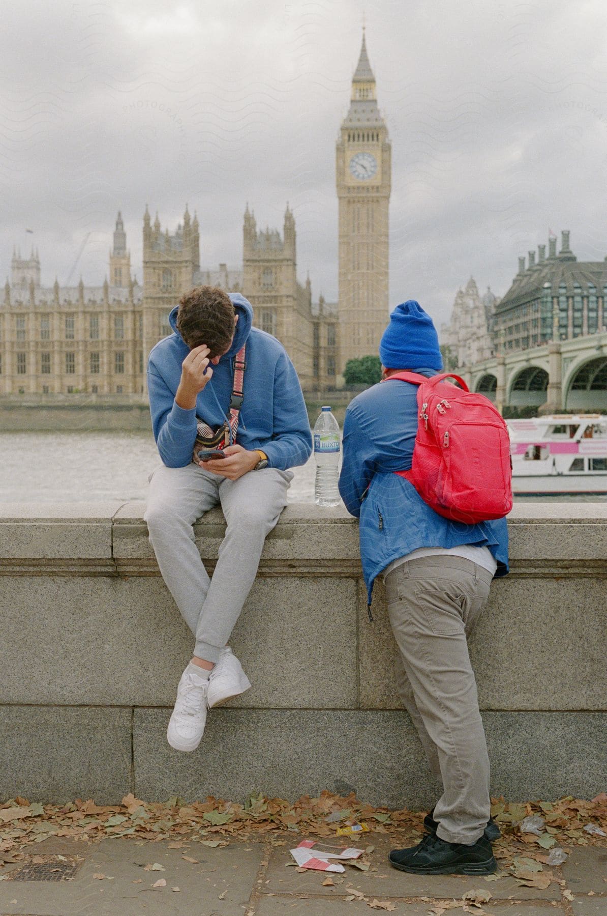 Two men in blue tracksuits sitting and leaning against a concrete structure by the edge of a lake.