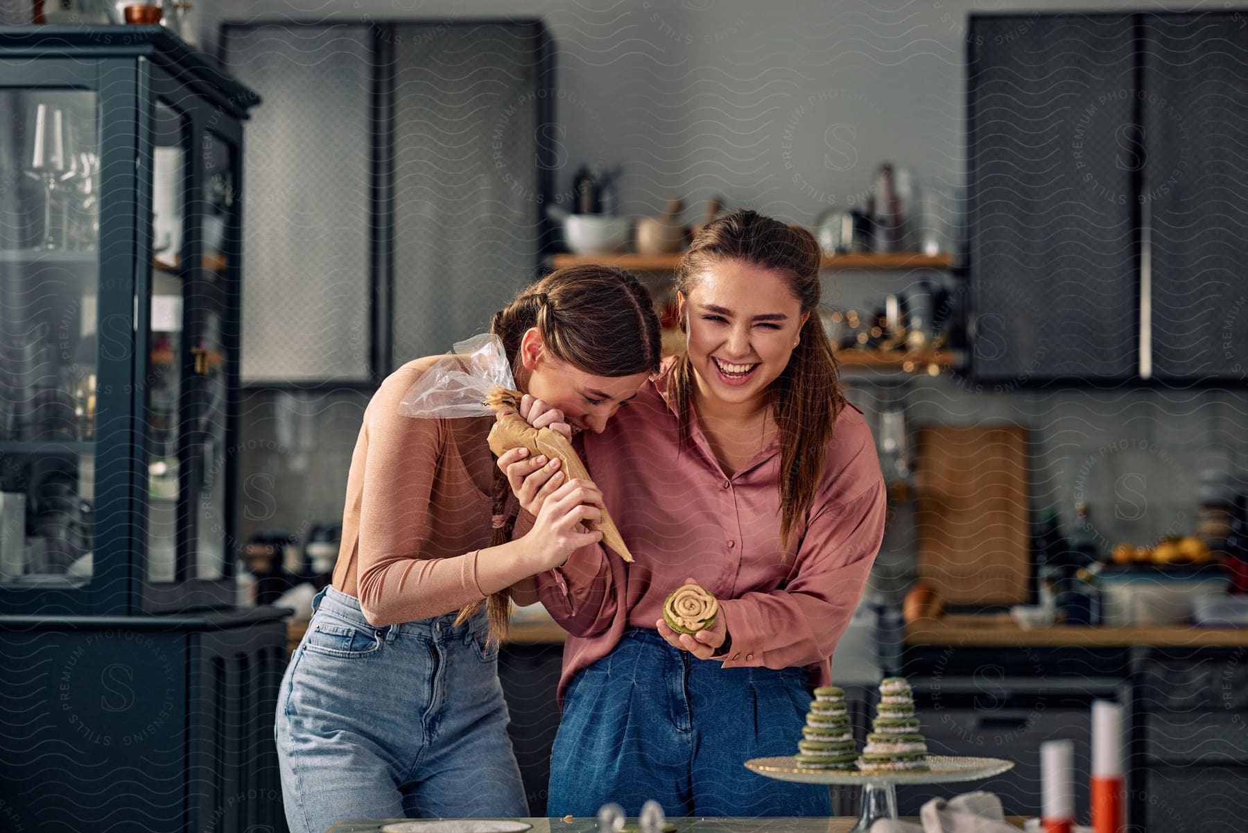 Two young women are piping frosting onto green sandwich cookies in a kitchen.