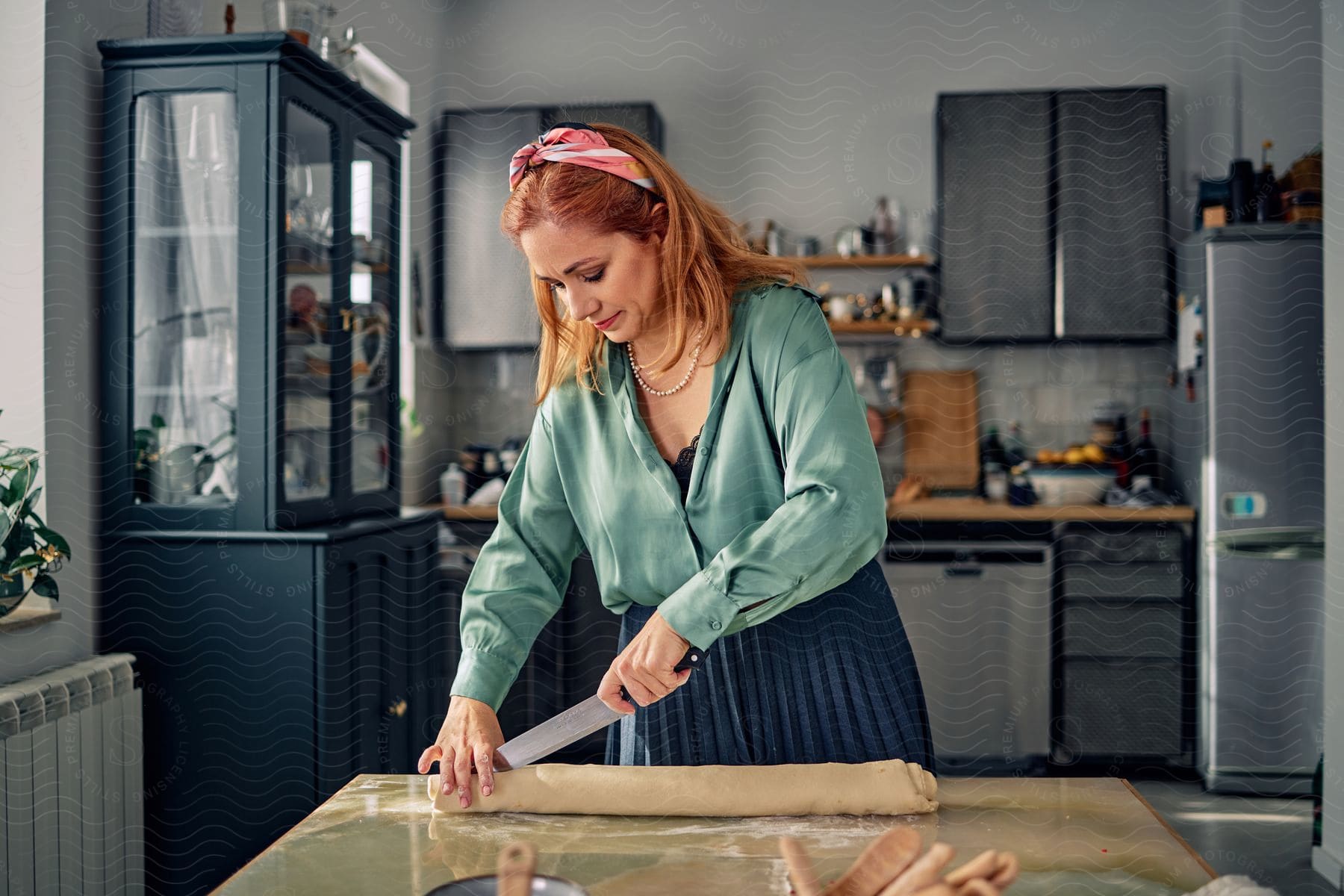 A woman wearing a turquoise shirt is using a knife to score a piece of dough on the dining table.