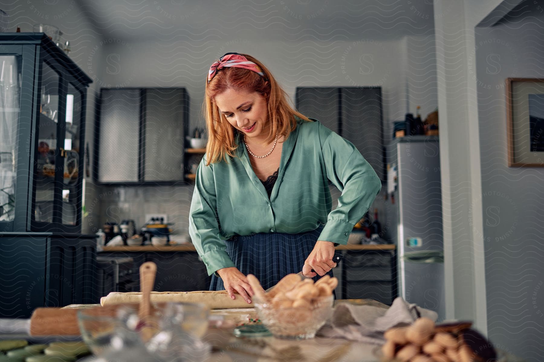 A mother with red hair, a green shirt, and a blue skirt cuts dough with a knife in the kitchen.