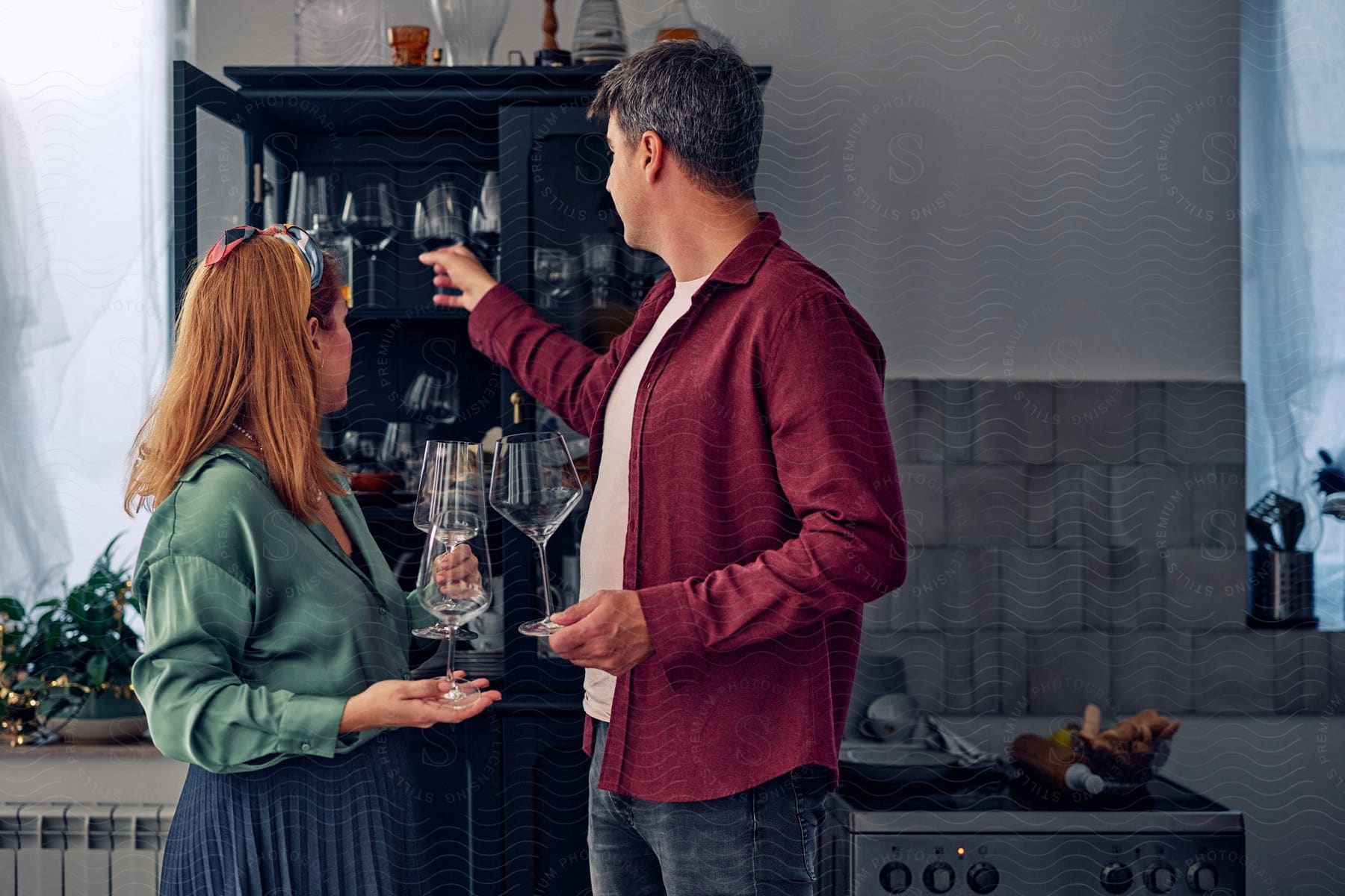 Couple taking crystal glasses from a black and rustic cabinet next to the stove in the kitchen.