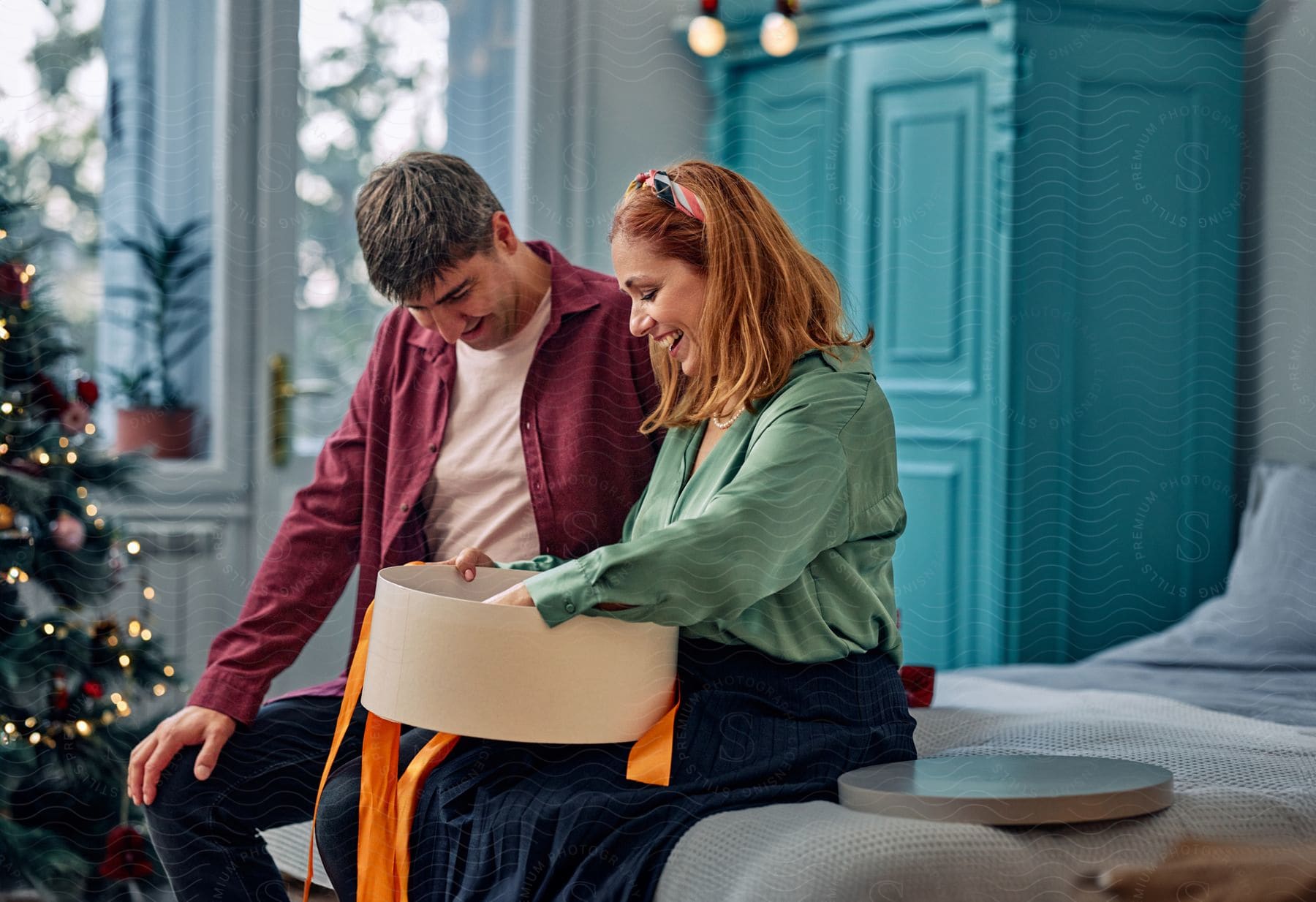 A Husband And His Wife Sitting On A Bed While Opening A Christmas Present