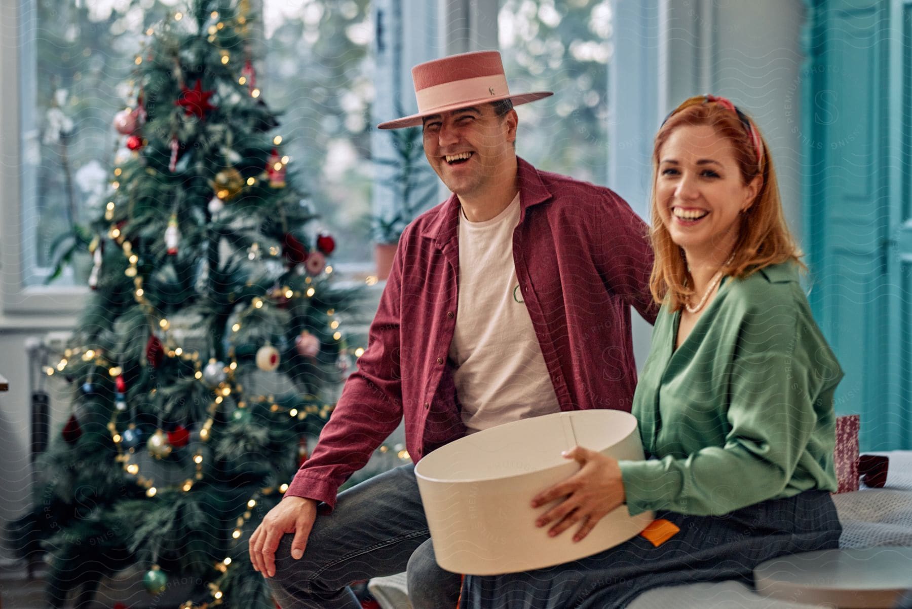 Smiling couple sitting, with the woman holding a gift box, and beside them a decorated Christmas tree.