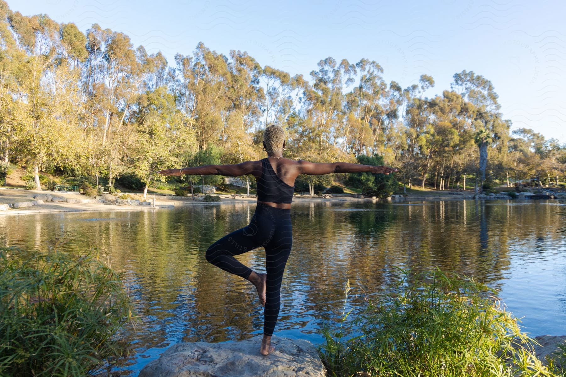 A woman balances on one leg while doing a yoga pose while outside near a lake.