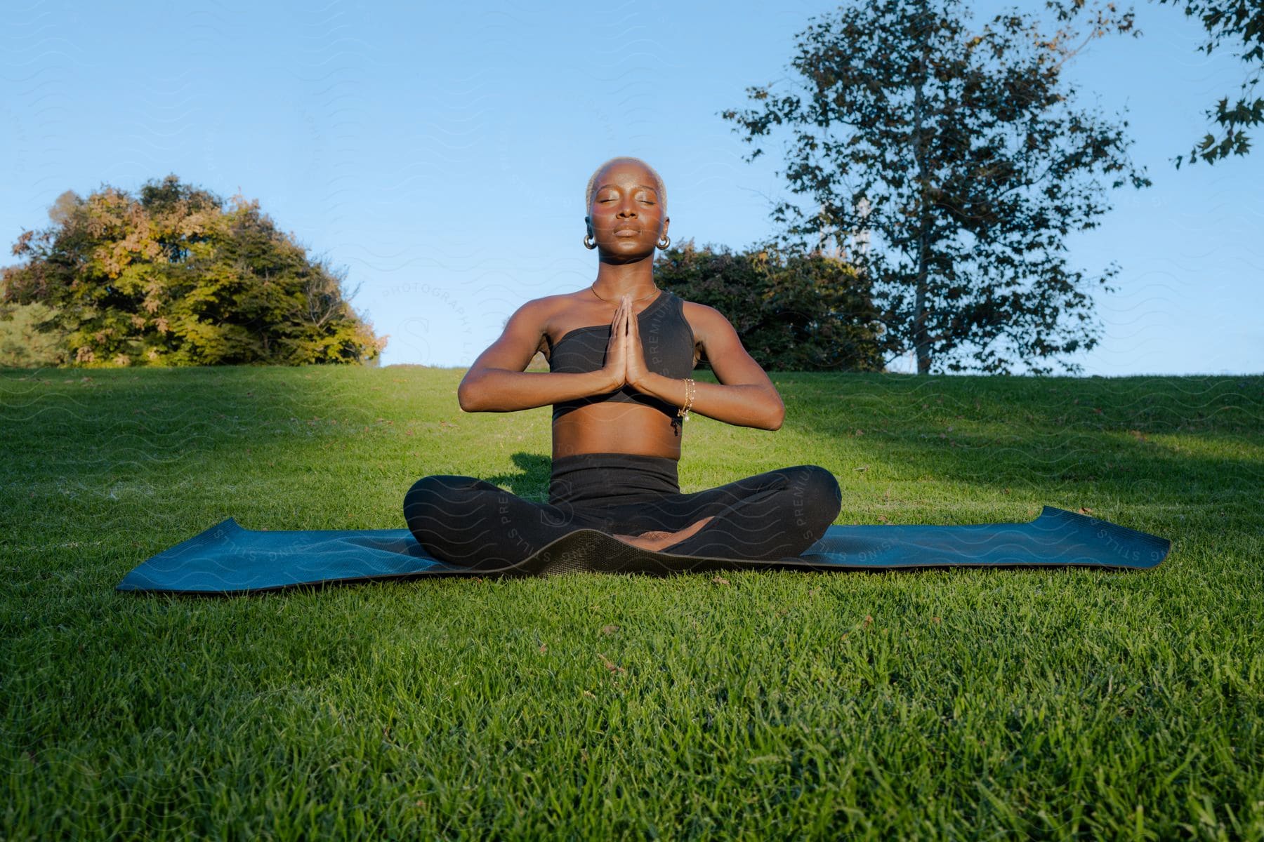 A woman sits on a mat in a field with her hands together and eyes closed as she meditates