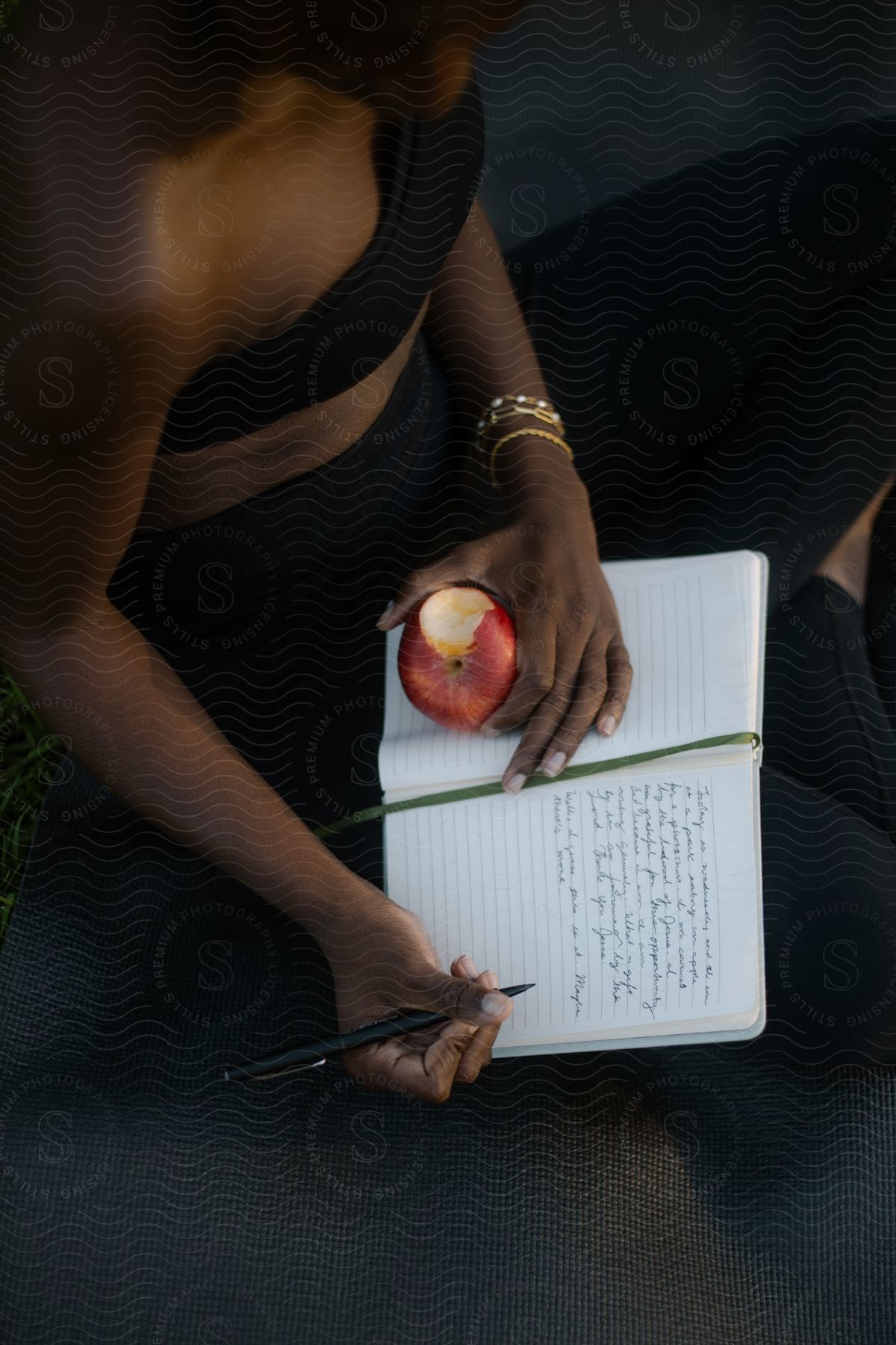A woman sits while writing in a lined notebook while holding an apple with a bite taken out of it.