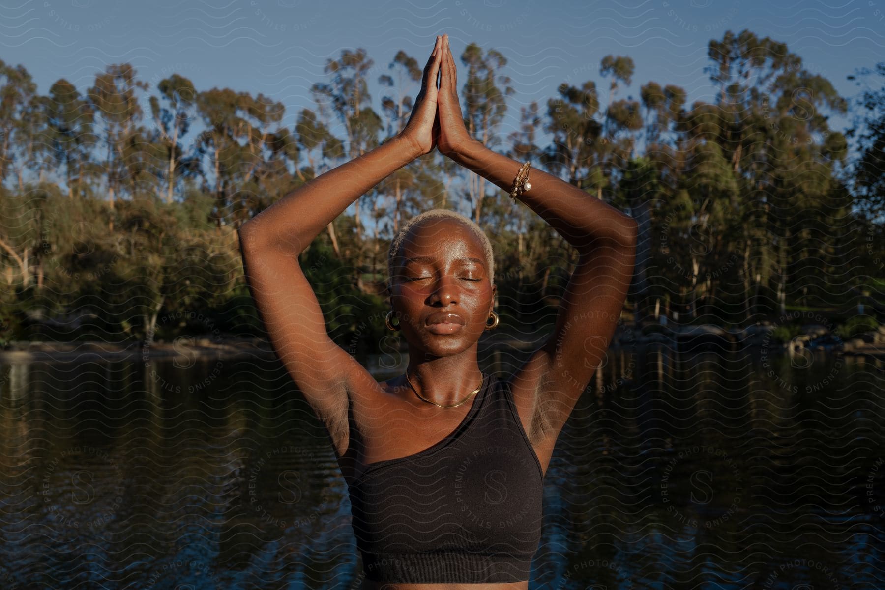 A woman holds her hands above her head while doing yoga outdoors near a lake.